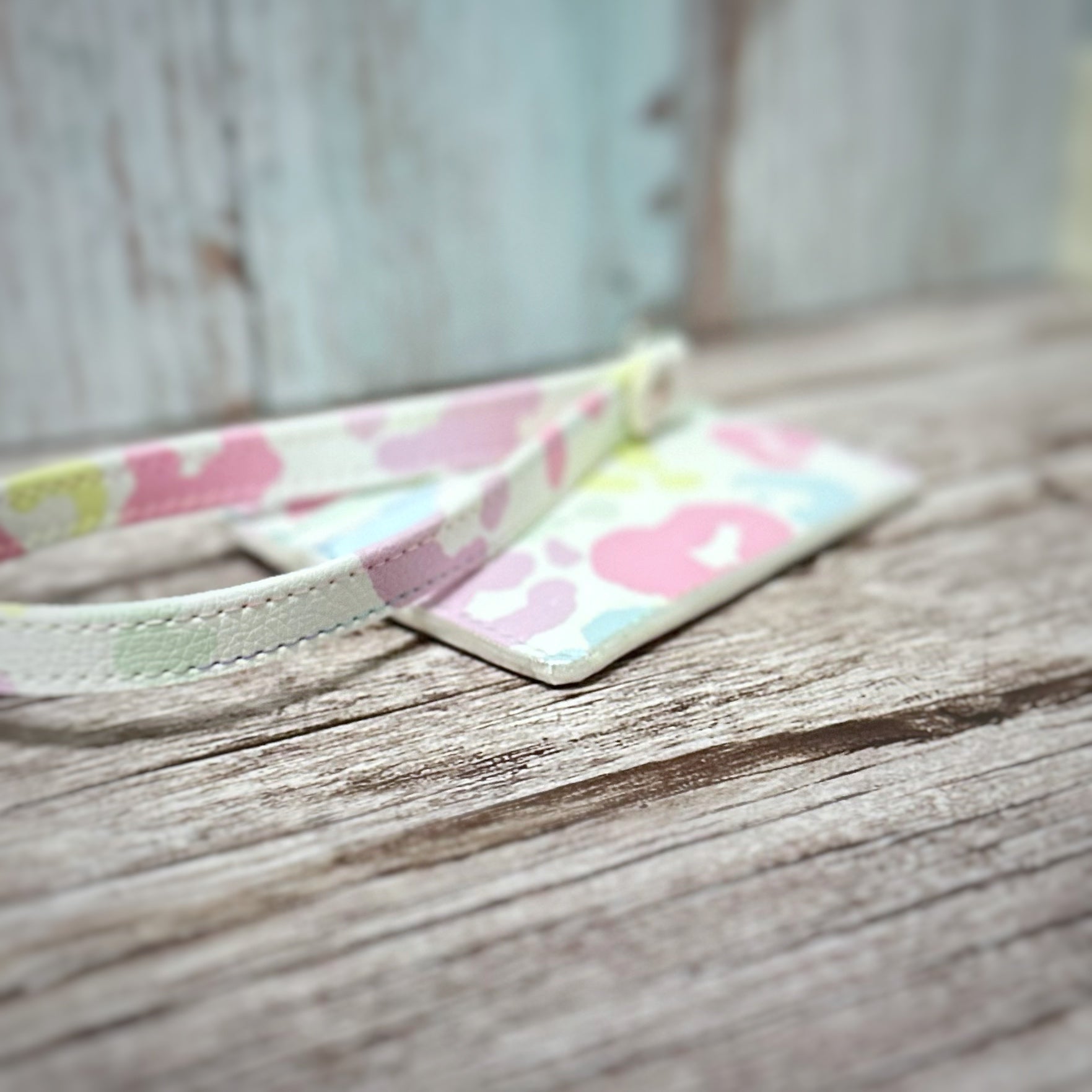 Close-up side view of the pastel cheetah print card holder, showcasing the shimmer white edge painting along the bottom seam. The detachable wristlet strap, also in matching pastel print, curves in the foreground. Displayed on rustic wood with a distressed pale teal and white background.