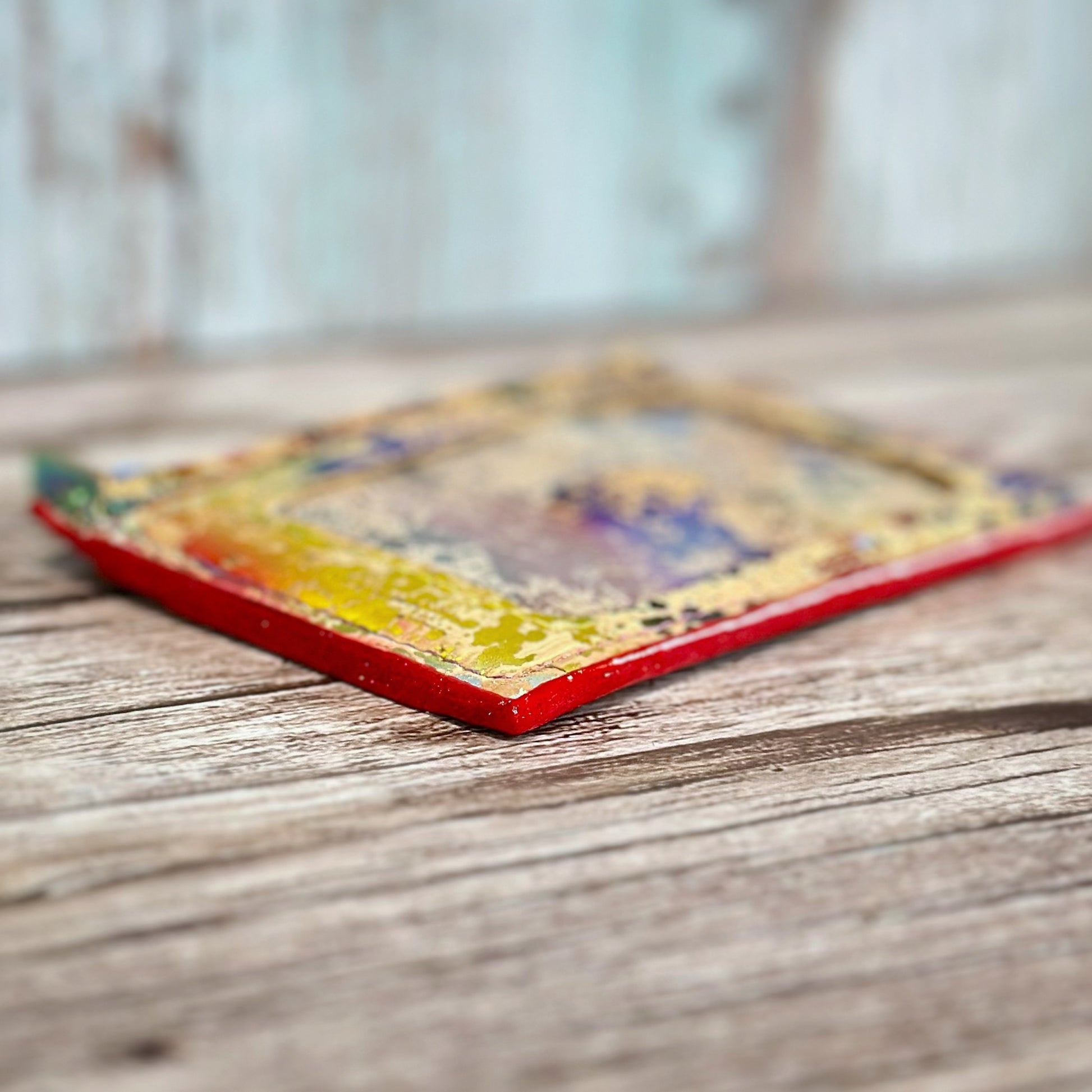 Side view of a handmade cork wallet lying flat, featuring red glitter edge coating and a vibrant multicolored paint-splatter pattern on a natural tan cork base. The wallet rests on a rustic wooden surface with a softly blurred background.