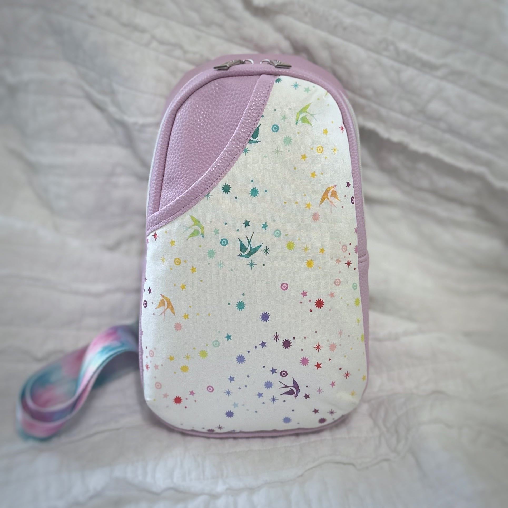 A handmade crossbody sling bag featuring a soft lavender vinyl body and a 3D cotton accent pocket on the front. The curved pocket panel showcases white fabric printed with scattered rainbow stars and swallows in flight. A coordinating pastel swirl crossbody strap peeks from the side. The bag is standing upright against a softly crinkled white fabric backdrop.