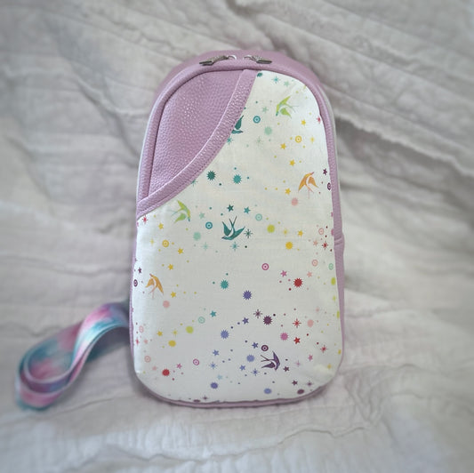 A handmade crossbody sling bag featuring a soft lavender vinyl body and a 3D cotton accent pocket on the front. The curved pocket panel showcases white fabric printed with scattered rainbow stars and swallows in flight. A coordinating pastel swirl crossbody strap peeks from the side. The bag is standing upright against a softly crinkled white fabric backdrop.