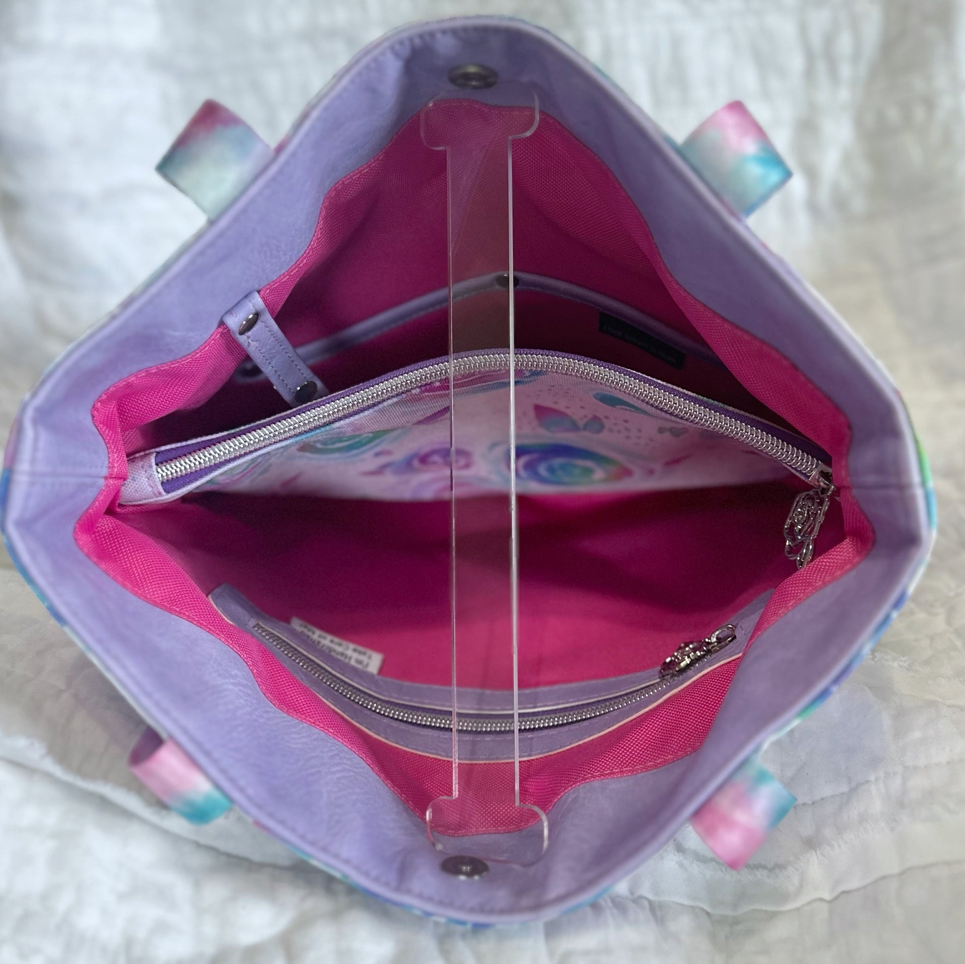 Interior view of a handmade tote bag showcasing a structured divider that doubles as a zippered pocket in the center. The inside lining is bright pink, with two slip pockets on one side and an additional zippered pocket on the opposite side. Lavender trim and floral print details coordinate with the exterior design. The bag is propped open to clearly display the interior layout.