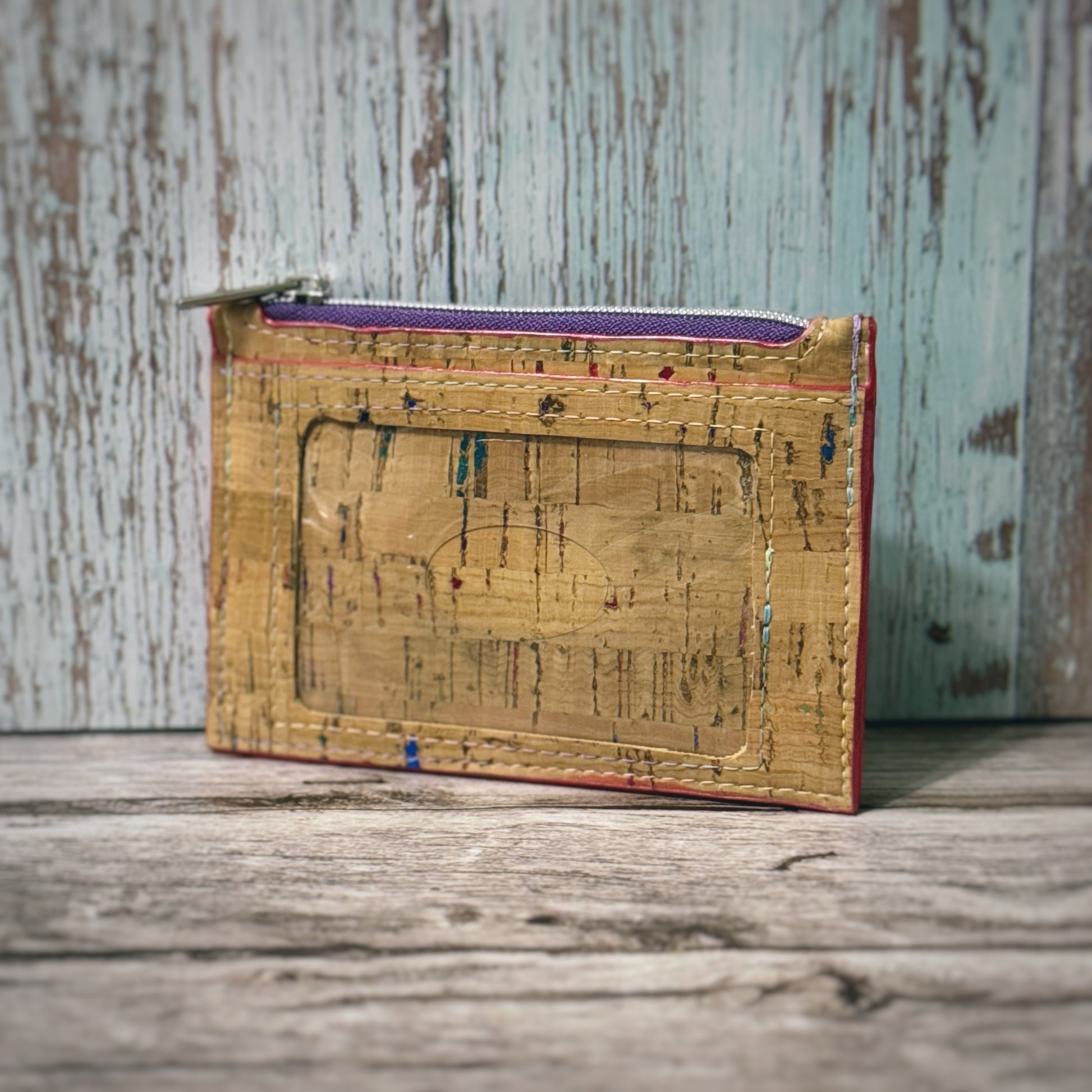 Handmade cork wallet with an ID window and top zipper closure. The cork material features a natural tan base with subtle multicolored flecks. The zipper is silver with a purple lining, and the wallet is displayed upright against a rustic wood background.