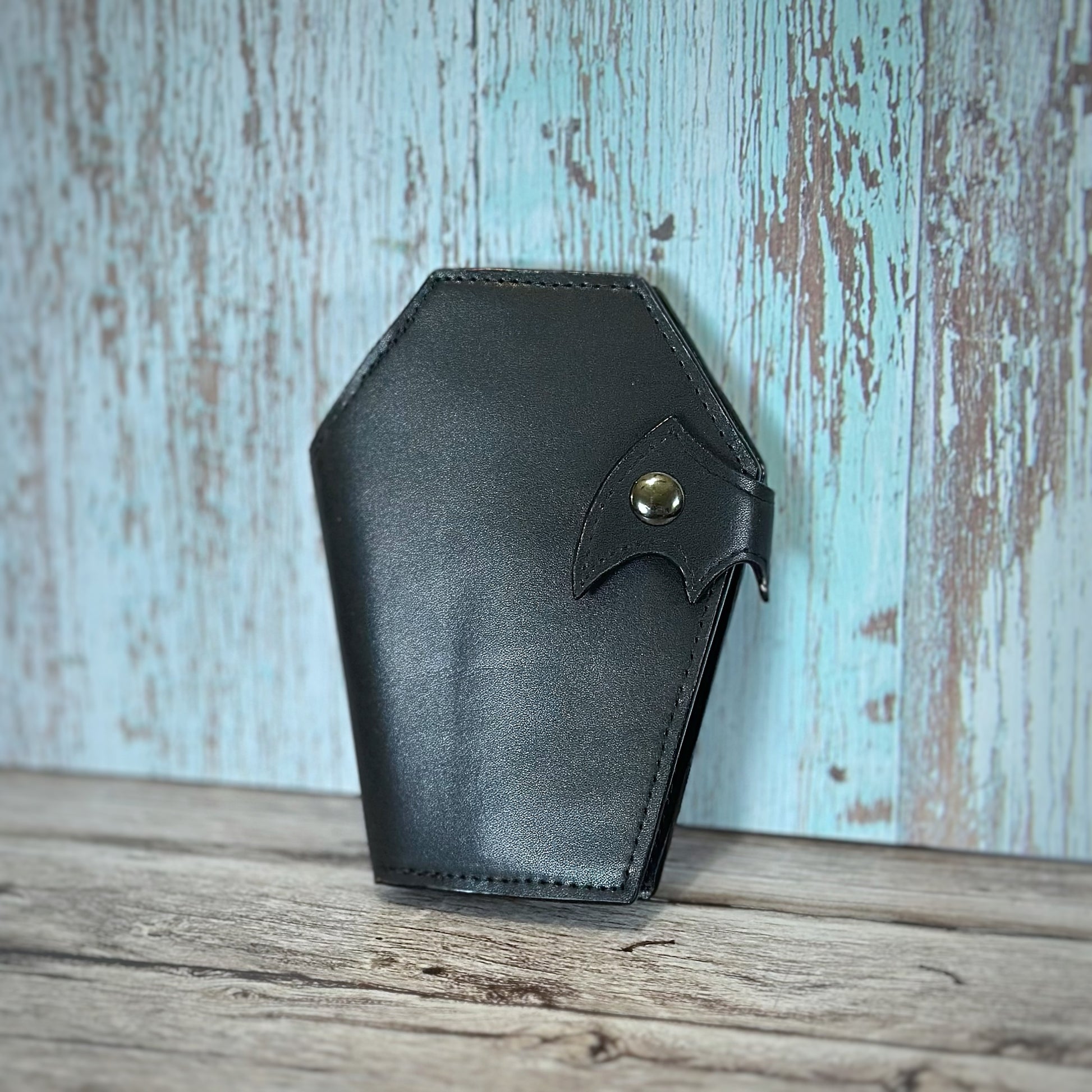Handmade coffin-shaped wallet crafted from smooth black vinyl with a subtle sheen. Features a bat wing flap closure secured by a gunmetal snap. Displayed upright on a rustic wood surface.