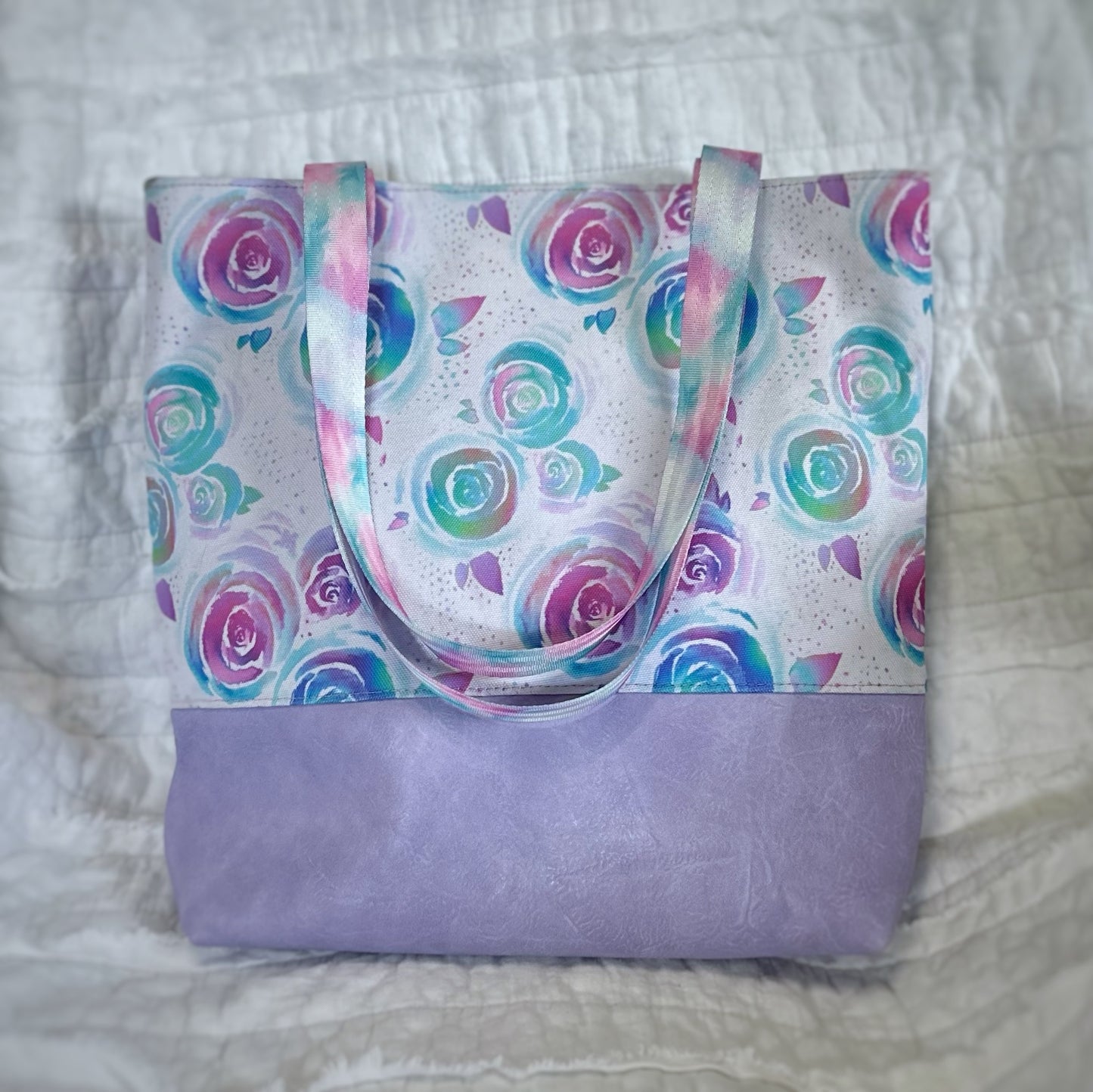 Handmade tote bag featuring a whimsical floral print in shades of teal, lavender, and pink on a white background. The bottom portion is made from soft lavender faux leather, with matching pastel rainbow handles. The bag is displayed upright against a quilted white backdrop.