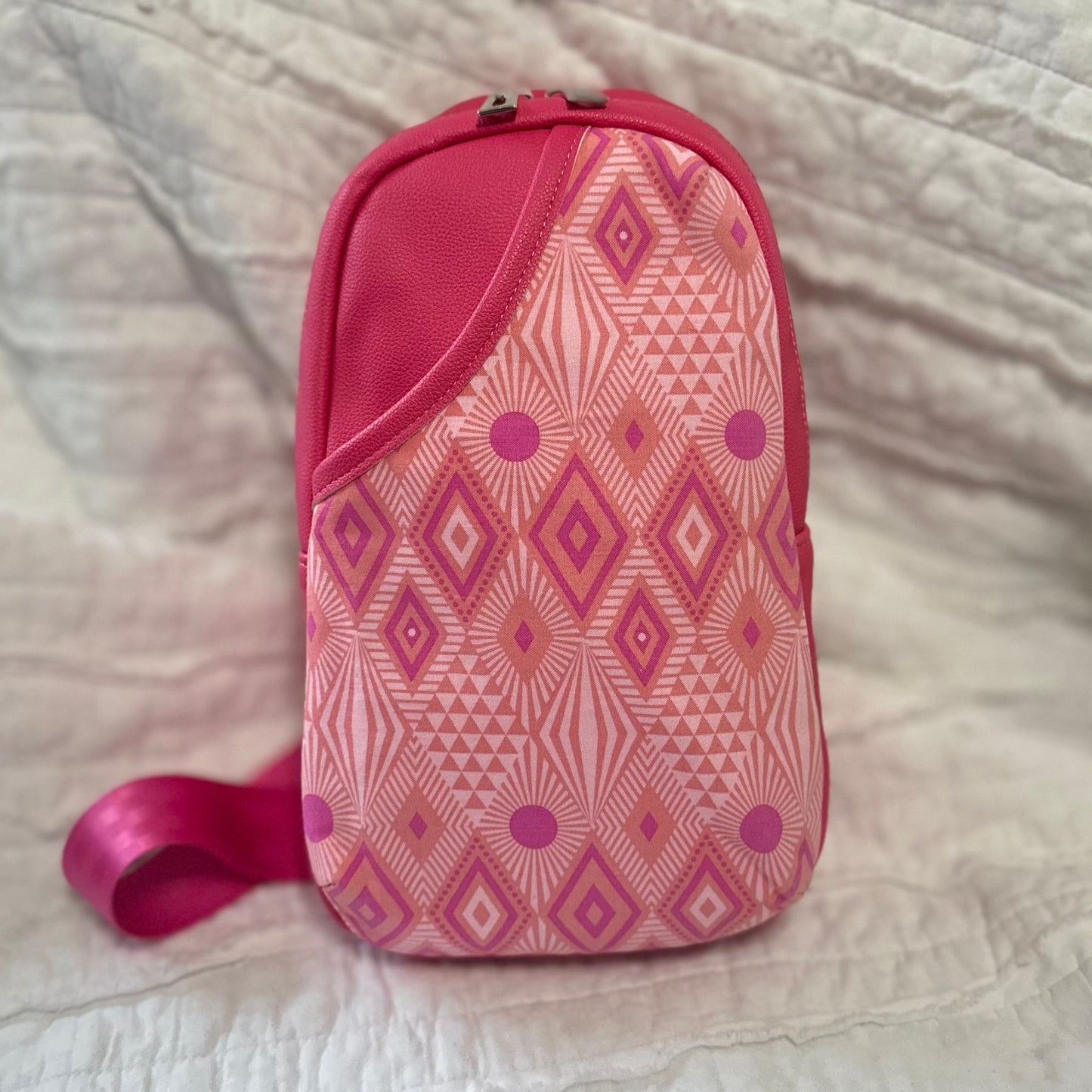 Front view of a handmade sling bag featuring a bold geometric cotton print in hot pink, coral, and magenta tones. The accent panel has a 3D curved shape and sits against a matching pink faux leather body. The bag includes a top zipper and a wide pink strap peeking out from the side.