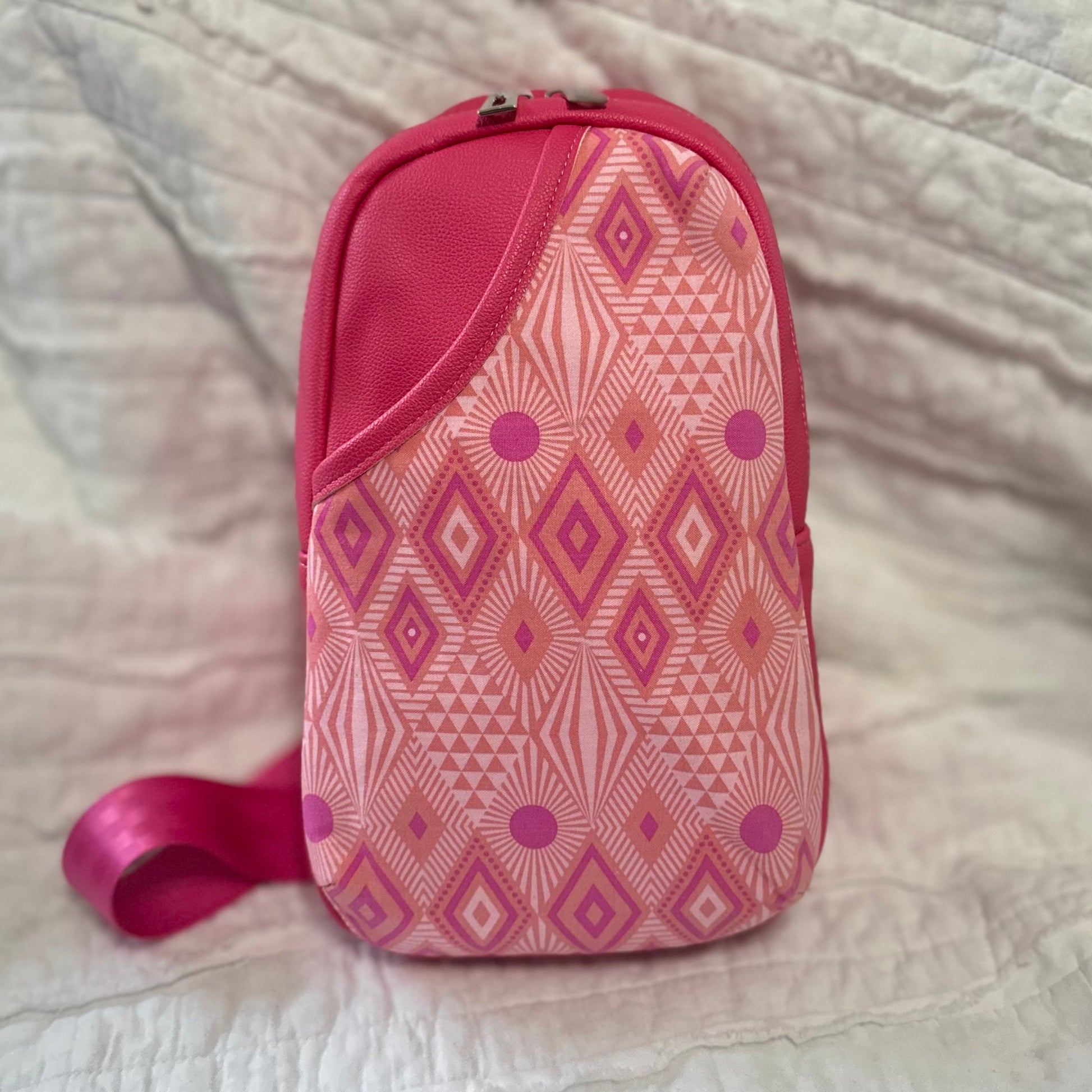 Front view of a handmade sling bag featuring a bold geometric cotton print in hot pink, coral, and magenta tones. The accent panel has a 3D curved shape and sits against a matching pink faux leather body. The bag includes a top zipper and a wide pink strap peeking out from the side.