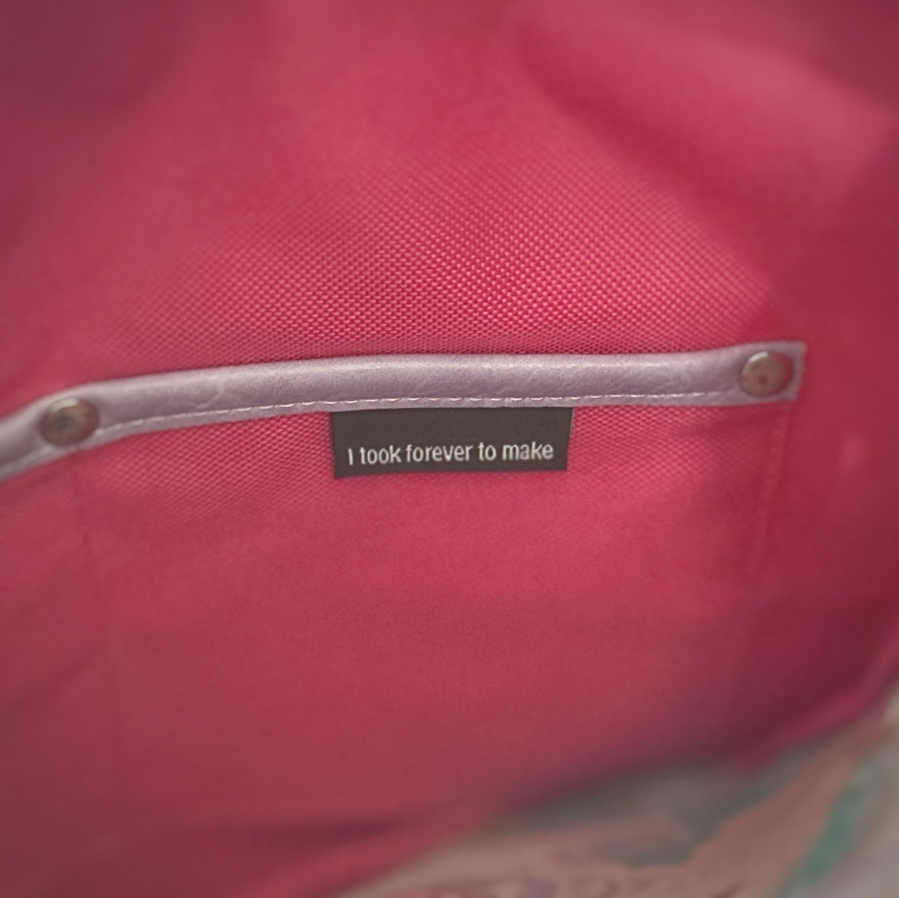 Close-up of the right slip pocket inside a handmade tote bag, made from bright pink fabric with lavender faux leather trim and metal rivets. A black fabric label is stitched into the pocket and reads, 'I took forever to make.'