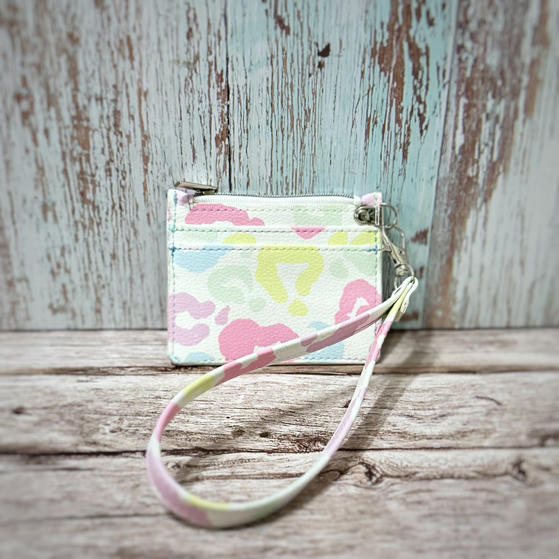 Handmade card holder with a detachable wristlet strap, crafted from pastel cheetah print faux leather in soft pink, mint, yellow, and lavender on a white background. Features two straight card slots, a top zipper closure, and silver hardware. The wristlet strap matches the print. Displayed upright on a rustic wooden surface with a distressed turquoise and white background.