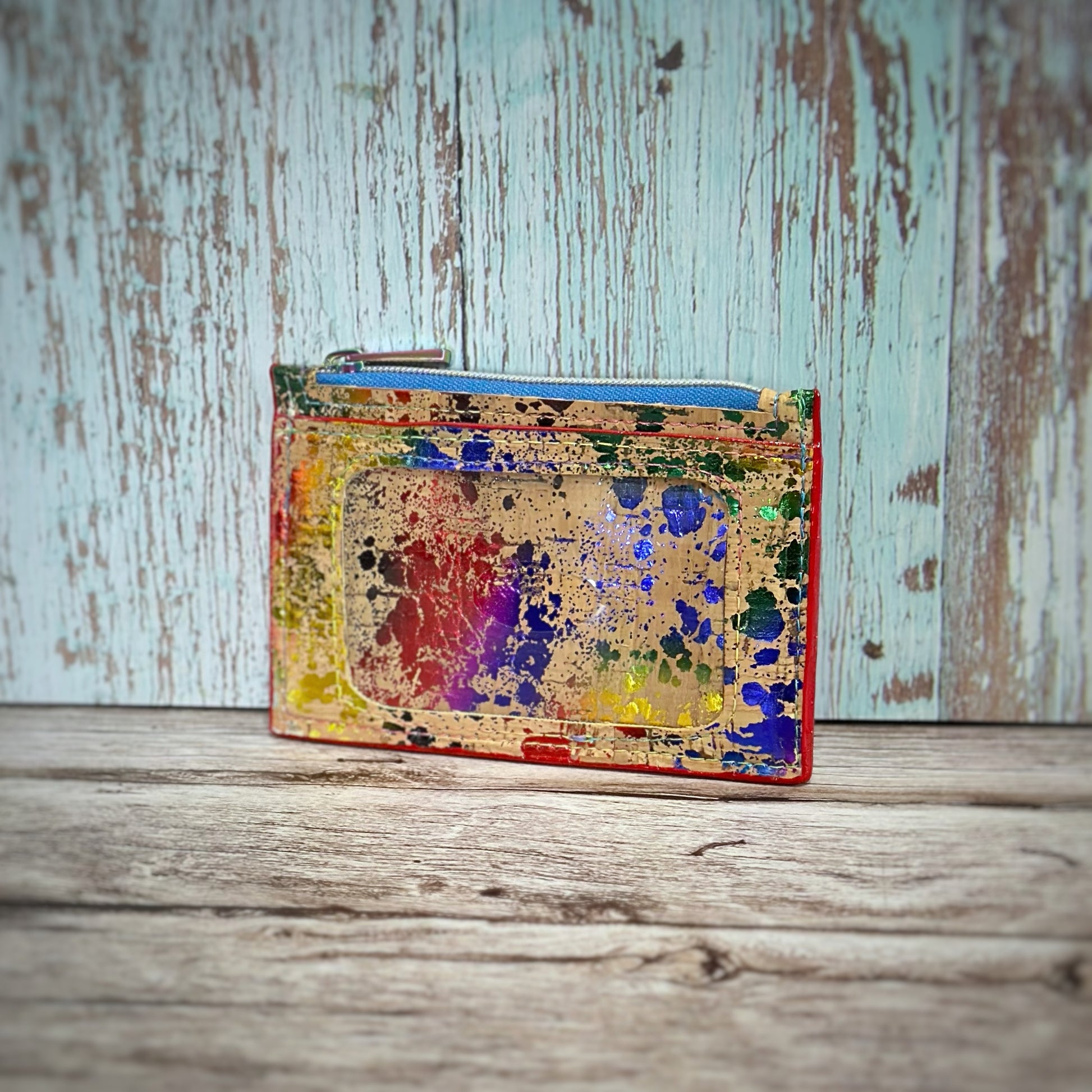Handmade cork wallet with an ID window and top zipper closure, featuring a vivid rainbow paint-splatter design across a natural cork base. The zipper is light blue with a rainbow pull, and the wallet is trimmed with red edges, displayed against a rustic wood backdrop