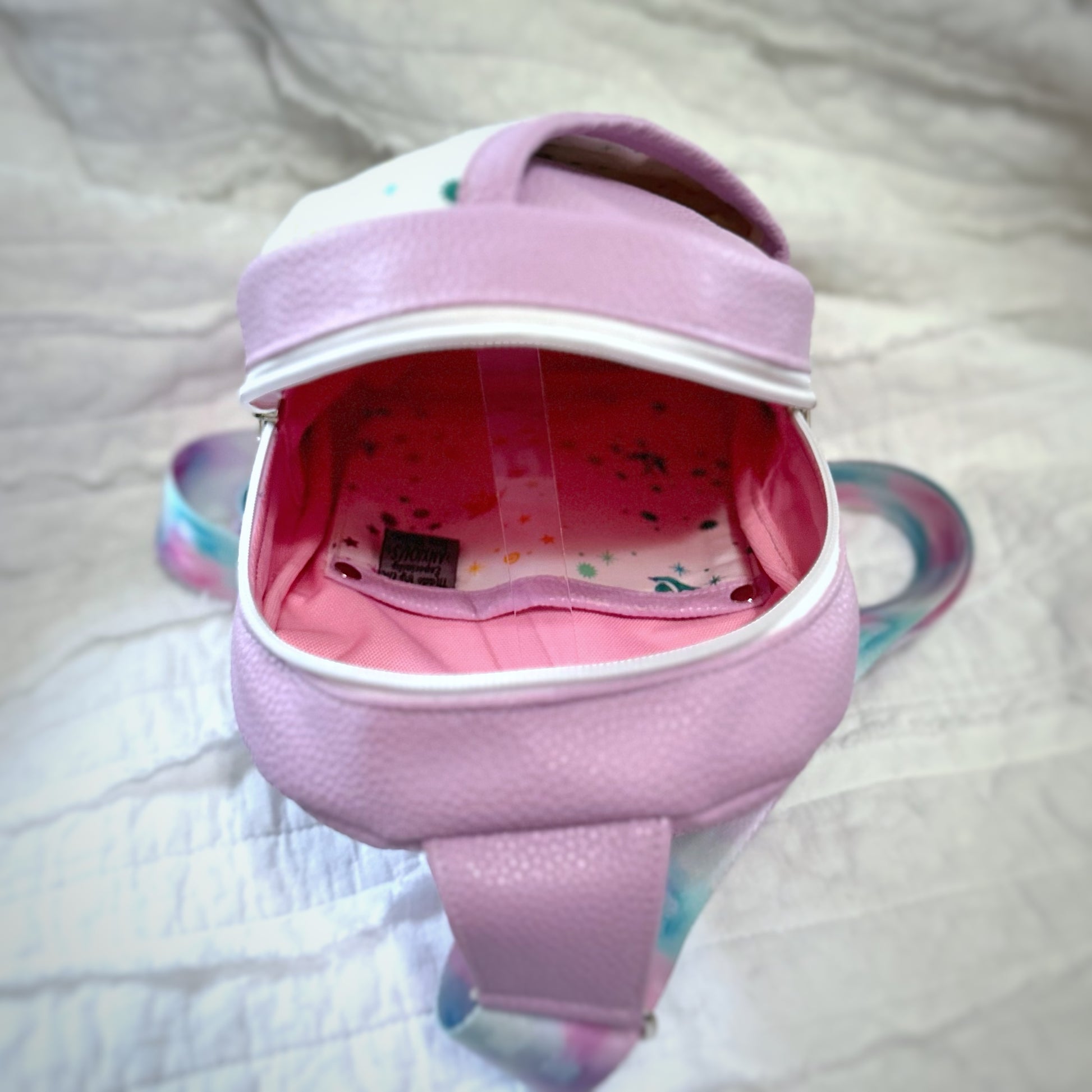 Interior view of a handmade sling bag, showing a coordinating slip pocket trimmed in lavender faux leather with silver rivet details at the corners. The inside fabric features a white background with scattered colorful stars and birds, matching the outer front pocket. The overall aesthetic is soft, whimsical, and neatly constructed.