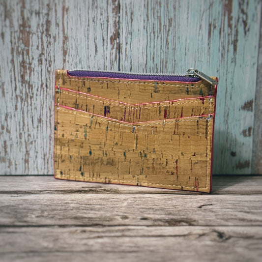Handmade cork wallet featuring two angled card slots on the front and a top zipper closure. The cork material is tan with subtle multicolored flecks, and the stitching is done in a contrasting red and white thread. The zipper has a silver pull and a purple lining, shown against a rustic wooden background
