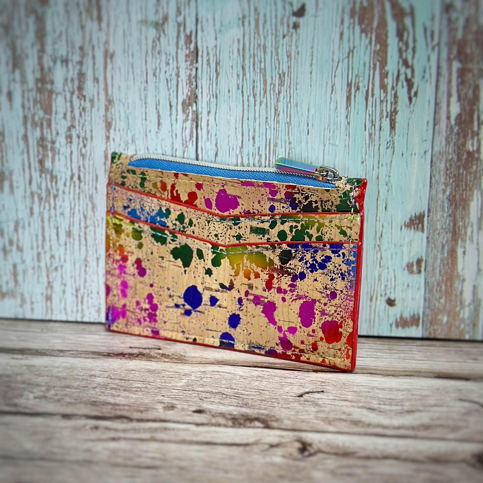 Handmade cork wallet with two angled front card slots and a top zipper closure, decorated in a bold, multicolored paint-splatter pattern on a natural tan cork background. The zipper is light blue with a silver pull, and the wallet is edged in red, shown standing upright against a rustic wooden surface.