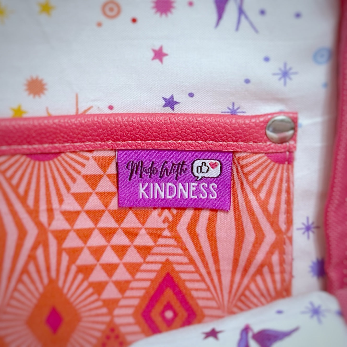 Close-up of the interior slip pocket on a sling bag, made from pink and coral geometric cotton with pink faux leather trim and a silver rivet. A bright purple woven tag is stitched to the pocket, reading 'Made with KINDNESS' in playful script and block letters. A thumbs-up icon with a heart is featured beside the text.