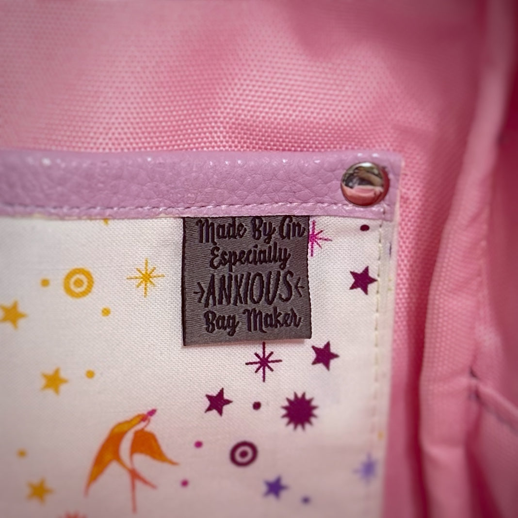 Close-up of the corner of a handmade bag’s interior slip pocket. The pocket is trimmed in lavender faux leather and secured with a shiny silver rivet. A woven label stitched onto the pocket reads: 'Made By An Especially Anxious Bag Maker.' The cotton fabric features a playful celestial print with scattered stars and a peach-colored bird, set against a pink background lining.