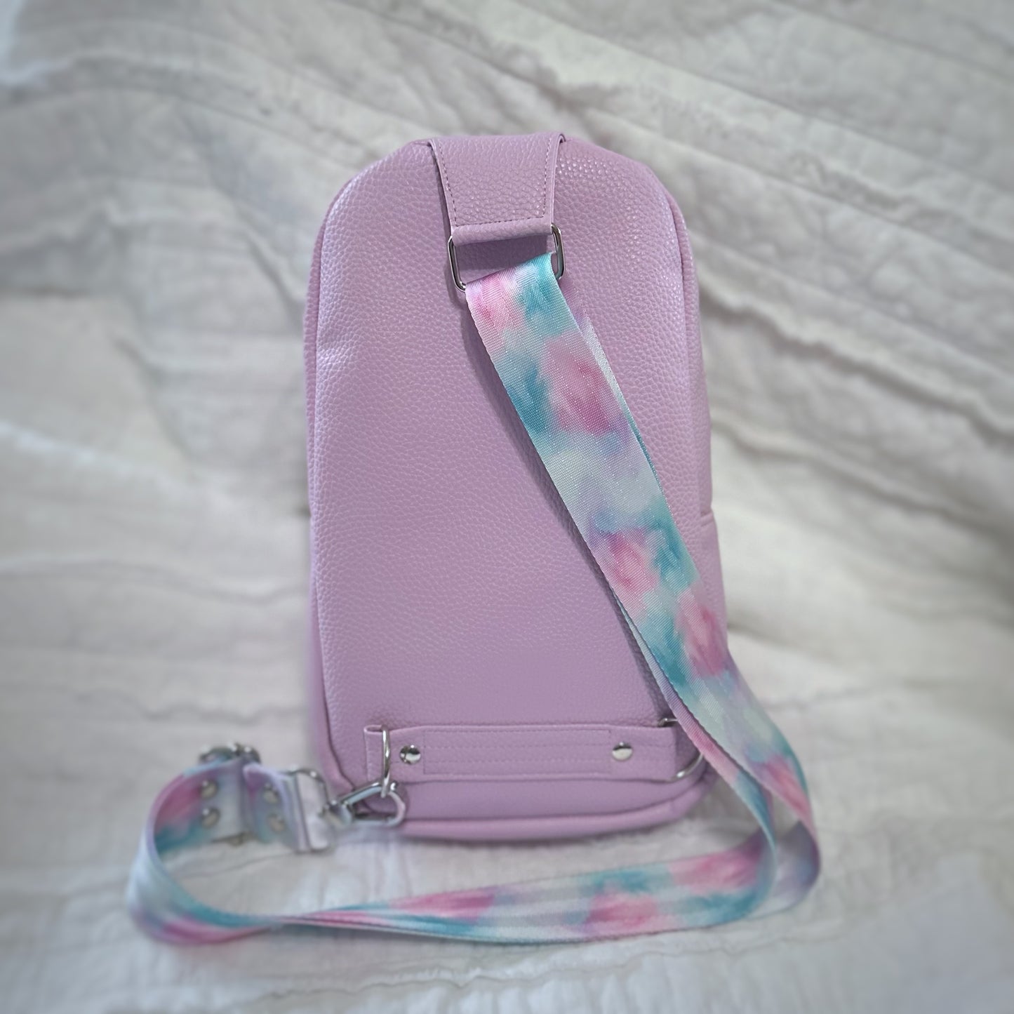 Back view of a handmade lavender vinyl sling bag, featuring a pastel swirl crossbody strap in pink, aqua, and purple hues. The strap attaches at the top and bottom of the bag with silver swivel hooks and D-rings. The backdrop is a softly crinkled white quilt, complementing the bag’s dreamy color palette.