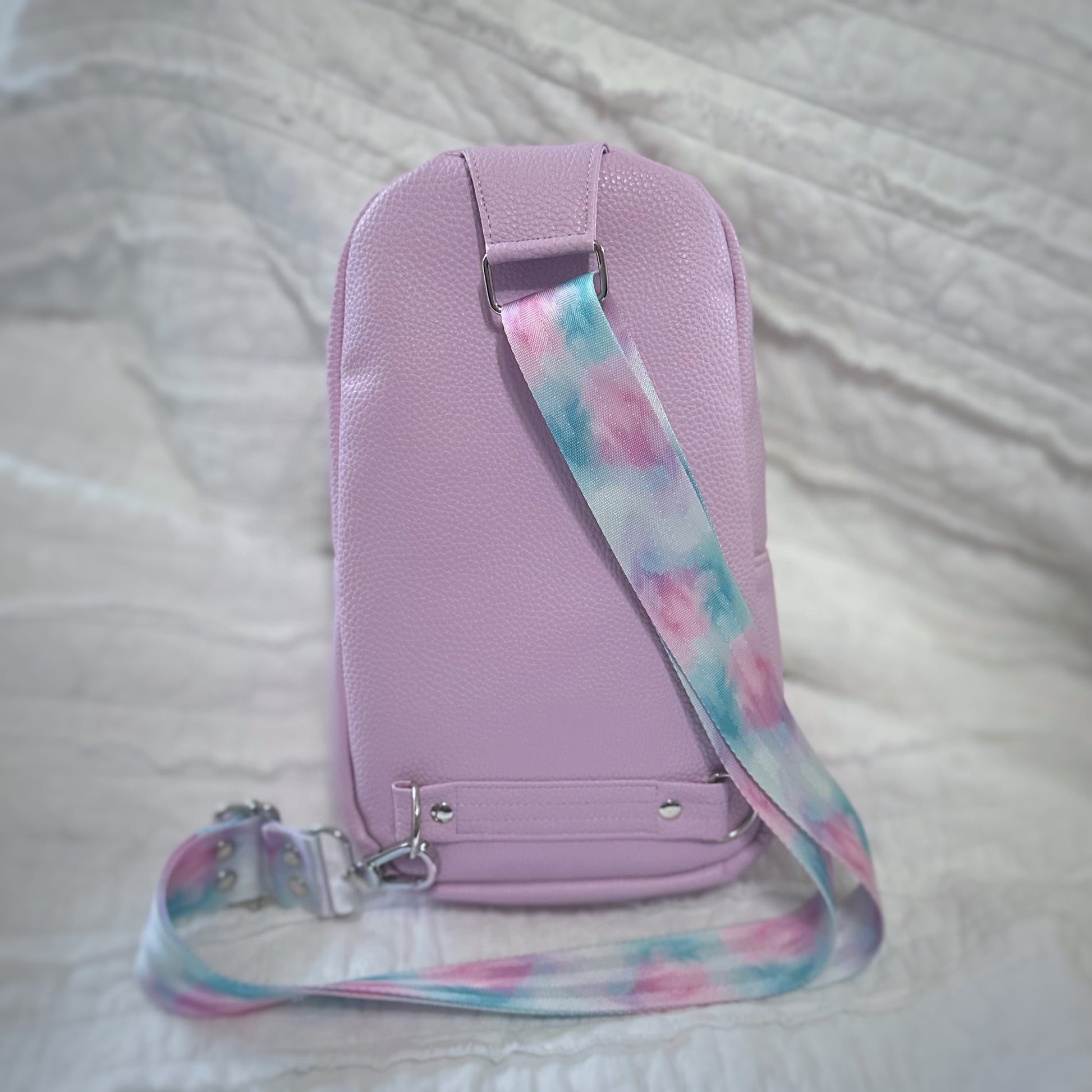 Back view of a handmade lavender vinyl sling bag, featuring a pastel swirl crossbody strap in pink, aqua, and purple hues. The strap attaches at the top and bottom of the bag with silver swivel hooks and D-rings. The backdrop is a softly crinkled white quilt, complementing the bag’s dreamy color palette.