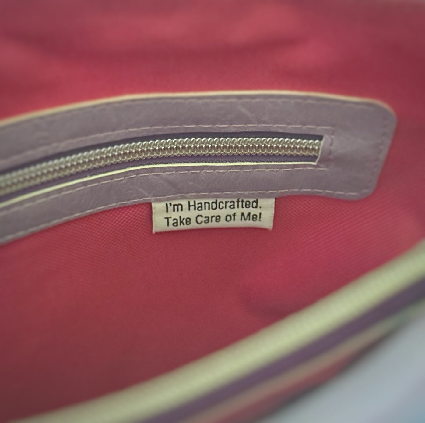 Close-up of the interior zipper pocket on a handmade tote bag. The zipper is trimmed with lavender faux leather and silver hardware. Below the zipper, a fabric tag reads, 'I'm Handcrafted. Take Care of Me!' against a bright pink fabric lining.