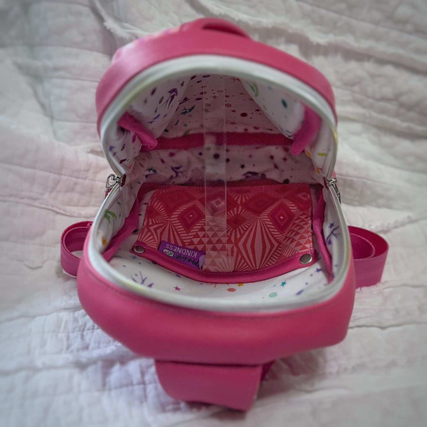 Interior view of a pink sling bag, showing a spacious compartment lined with white cotton printed in rainbow-colored stars and birds. Inside is a slip pocket made from matching pink geometric cotton, secured with silver rivets and trimmed in coordinating pink faux leather. The bag is unzipped fully to display the details clearly.