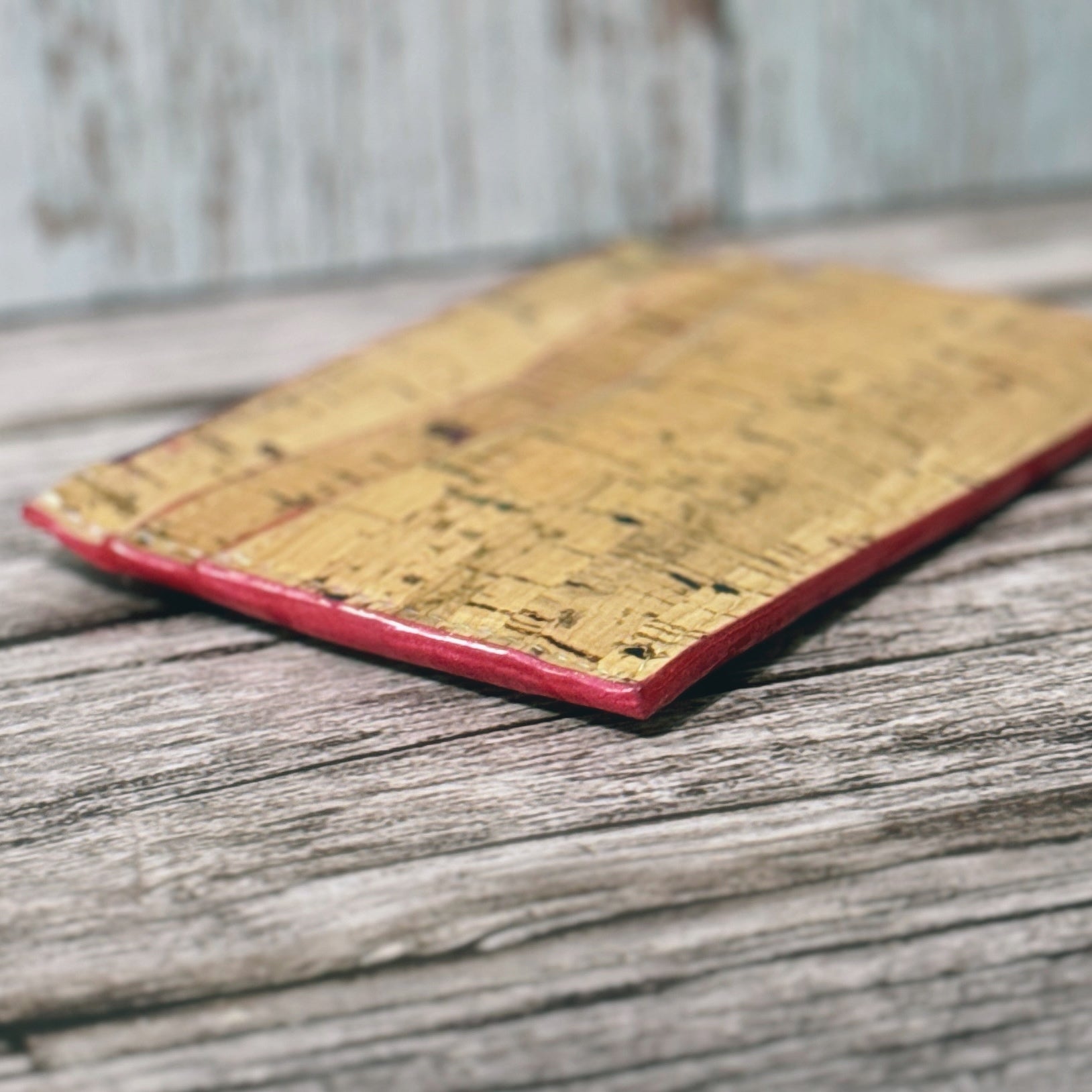 Close-up angled view of a slim handmade cork wallet lying flat on a rustic wood surface. The wallet features natural tan cork with multicolored flecks and neatly finished hot pink edge coating for a polished, professional look.