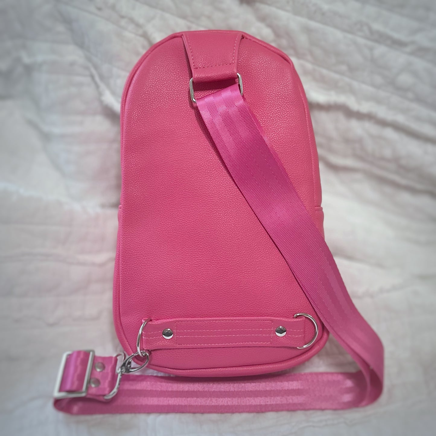 Back view of a handmade sling bag in vibrant pink faux leather with a matching pink webbing crossbody strap. The strap is connected at the bottom with silver hardware and reinforced with a riveted faux leather strip for durability.
