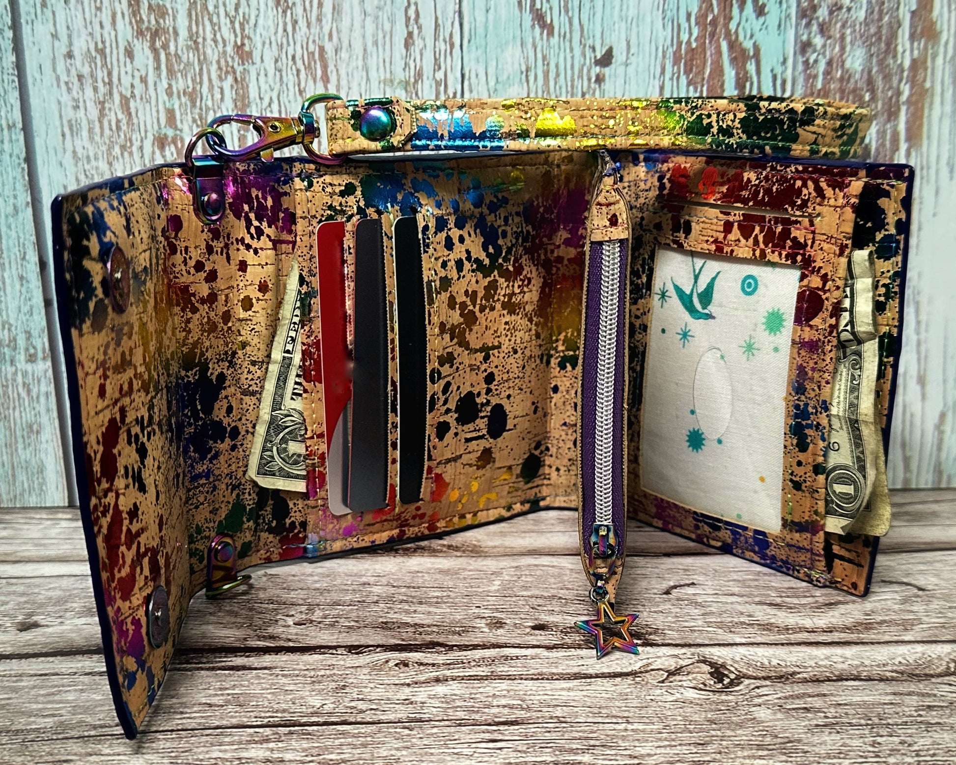 Interior view of a handmade wallet with a rainbow paint-splatter cork design. The left panel features three horizontal card slots with cards peeking out of each and a folded dollar bill in the cash pocket behind. The center includes a zipper pocket with a rainbow star charm pull. The right panel shows a clear ID window and a second cash pocket behind it, also with a folded dollar bill visible. The wallet is propped open with the wristlet strap atop on a wooden surface with a distressed wood backdrop.
