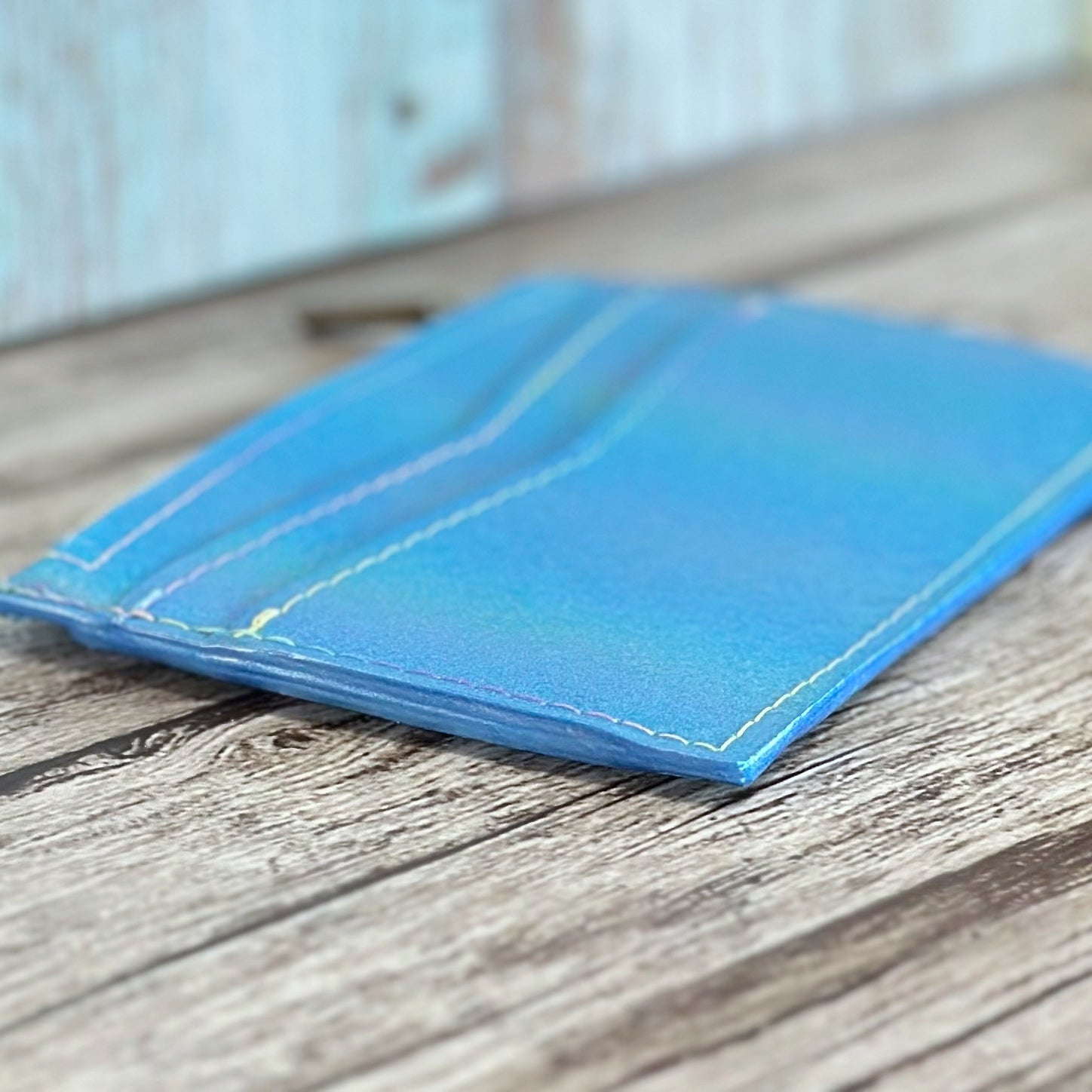 Angled close-up of a handmade wallet lying flat, made from smooth iridescent blue vinyl with a soft color shift and subtle sheen. The wallet features front card slots, clean white topstitching, and matching shimmer blue edge paint for a polished finish. Displayed on a rustic wooden surface with a softly blurred background