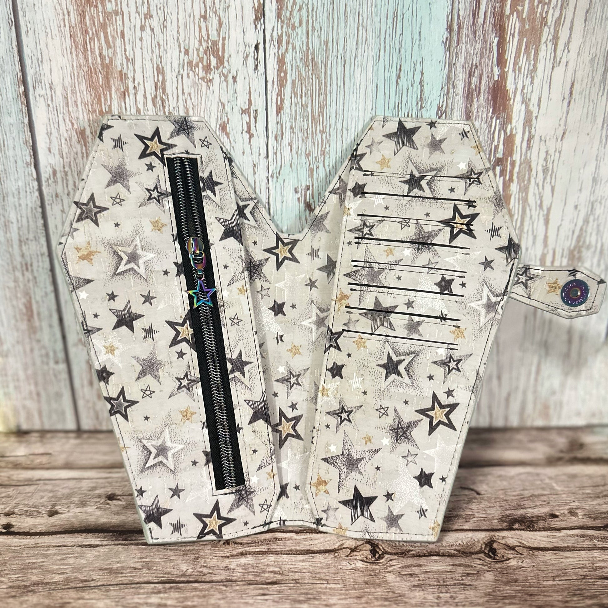 Interior view of a tall, handmade coffin-shaped wallet crafted from black, gold, and white star-print cork fabric. The left panel features a black zipper pocket with a rainbow-finish star-shaped zipper pull. The right panel includes 8 horizontal card slots, with a cash slot hidden behind each side. The coffin-shaped snap tab closure with rainbow snap is visible on the right.