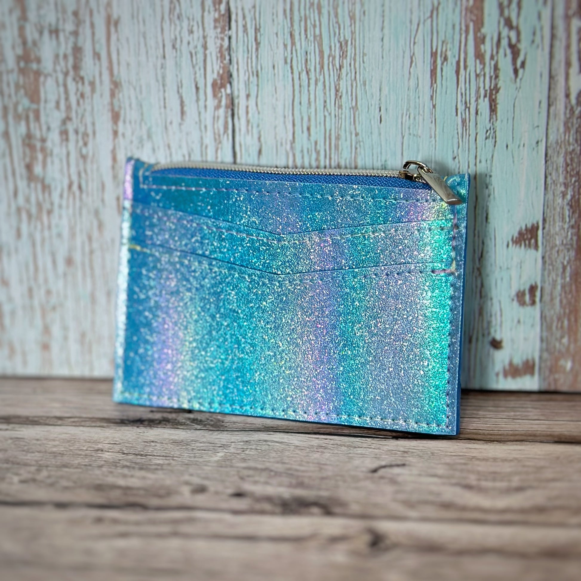 Handmade wallet made from iridescent blue glitter vinyl, featuring two angled front card slots and a top zipper closure. The shimmering surface reflects shades of aqua, lavender, and silver. The zipper is silver with a matching pull, and the wallet is photographed upright against a rustic wooden backdrop