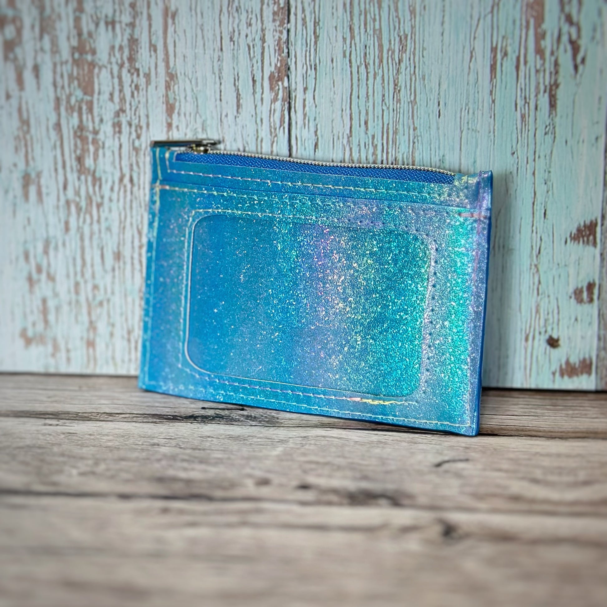 Handmade wallet crafted from iridescent blue glitter vinyl with a clear ID window and top zipper closure. The shimmering material shifts with hints of aqua, lavender, and silver in the light. The zipper is blue with a silver pull, and the wallet is displayed against a weathered wood background