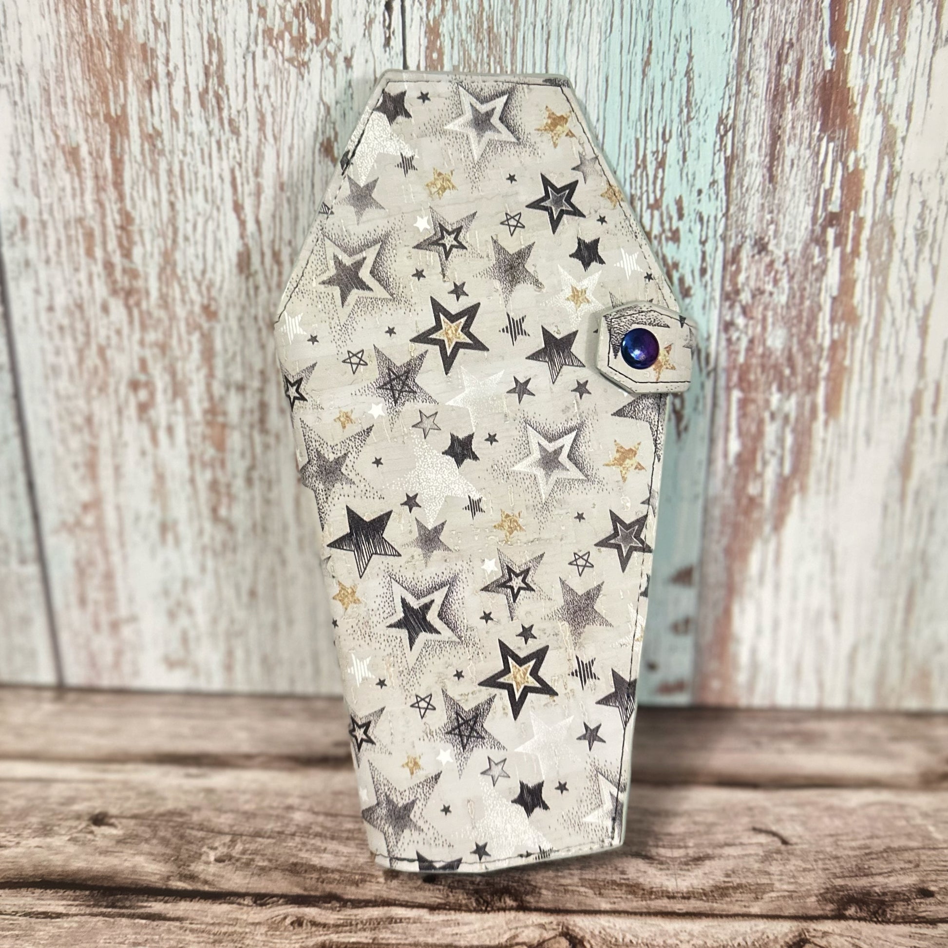 Front view of a handmade tall coffin-shaped wallet made from star-patterned cork fabric in shades of black, white, and gold on a light background. The wallet features a coordinating coffin-shaped snap tab with a rainbow-finish snap closure.
