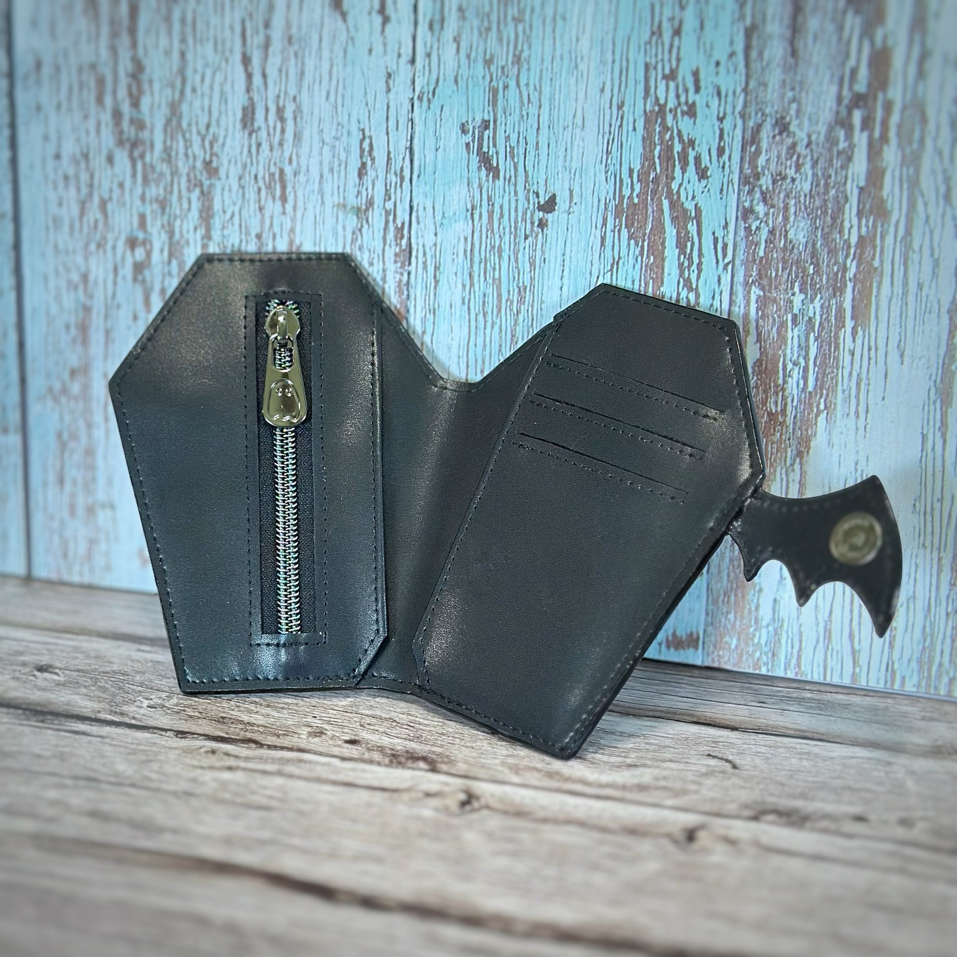 Interior of a handmade coffin-shaped wallet made from smooth black vinyl. Features three horizontal card slots, a vertical zipper pocket with gunmetal hardware, and a full-length cash slot hidden behind both sections. The bat wing flap closure is open and secured with a matching gunmetal snap. Displayed open on a rustic wooden surface.