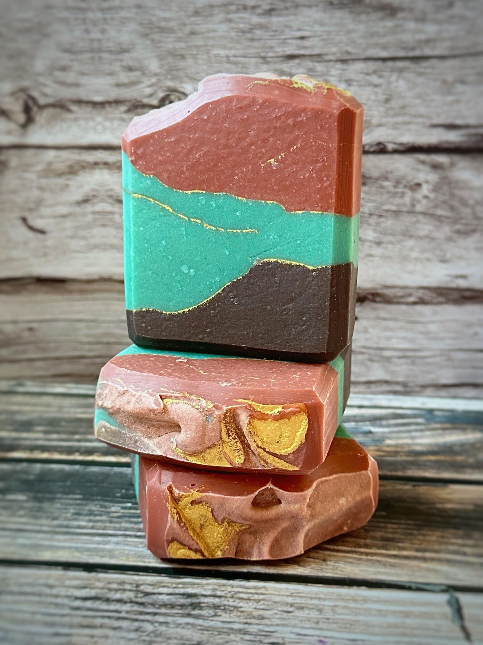 Three handcrafted soap bars stacked on a rustic wooden surface. Each bar showcases rich layers of oxidized copper tones - weathered teal, deep rust, and aged bronze - intertwined with shimmering veins of metallic gold. The top bar features a softly sculpted top edge, while the lower bars reveal swirled golden accents that evoke the organic beauty of a well-aged patina. A muted wood plank backdrop enhances the antique, industrial charm of the design.