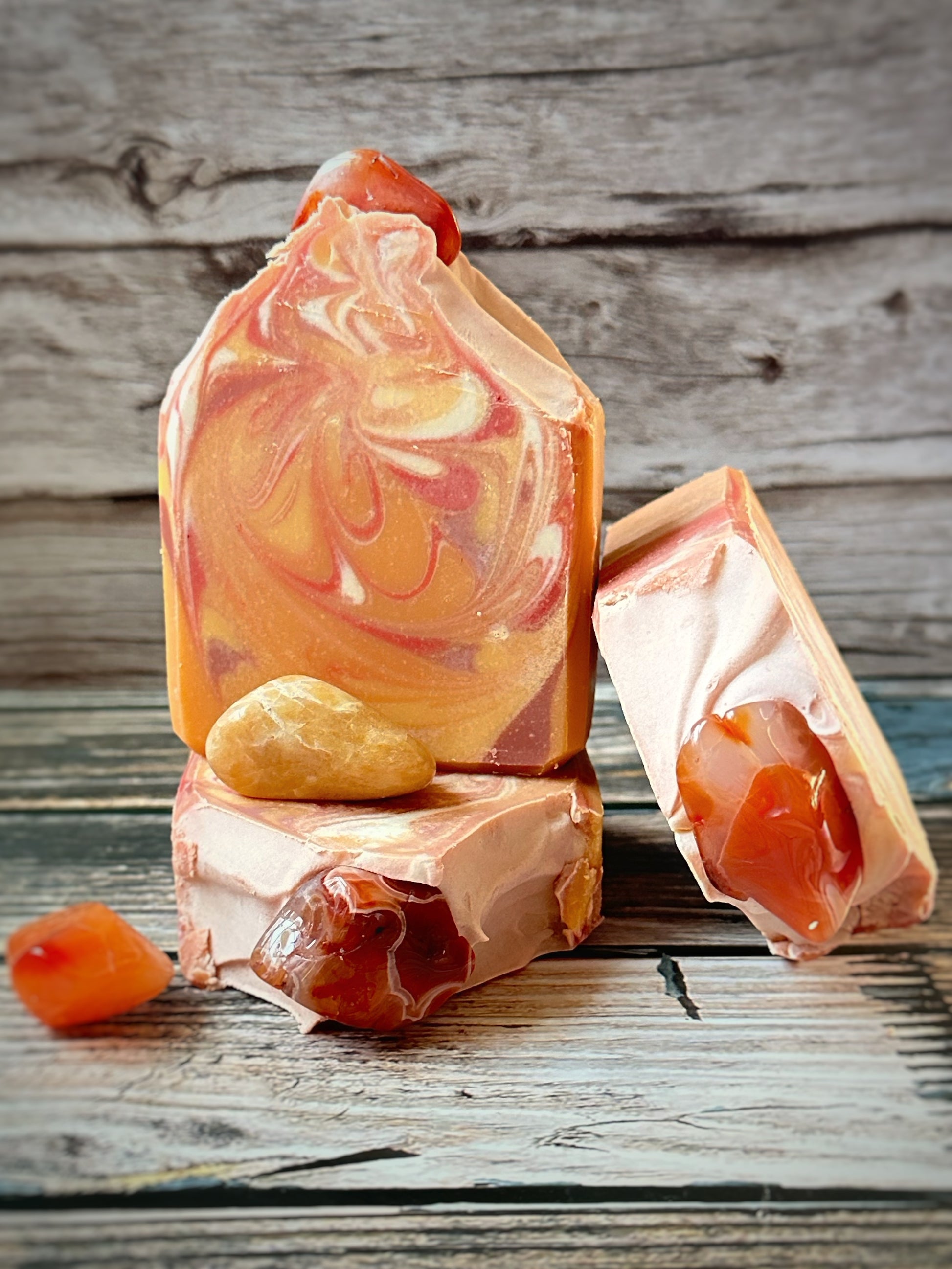Three orange and coral-toned artisan soap bars arranged on a wood surface, featuring bold swirls of peach, rust, and cream. The soaps are embedded with real carnelian stones. Additional polished carnelian stones are placed around the base for accent.  The background is rustic wooden paneling.