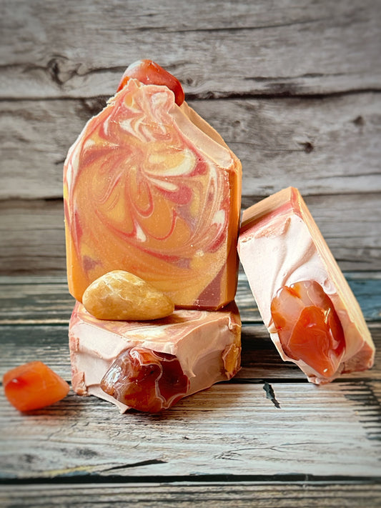 Three orange and coral-toned artisan soap bars arranged on a wood surface, featuring bold swirls of peach, rust, and cream. The soaps are embedded with real carnelian stones. Additional polished carnelian stones are placed around the base for accent.  The background is rustic wooden paneling.