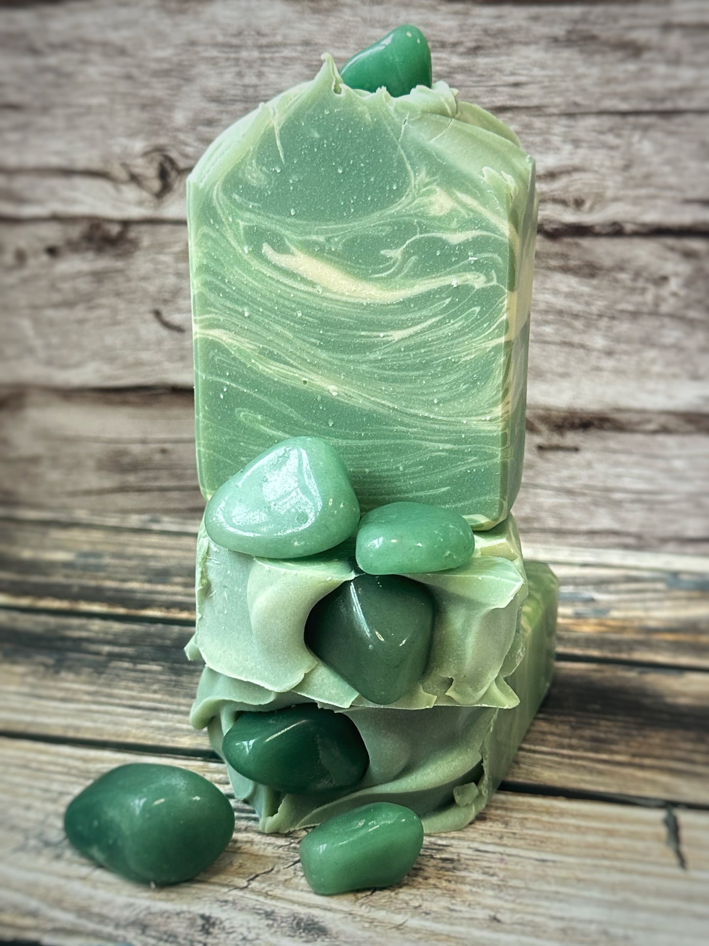 Three green artisan soap bars stacked with real green aventurine stones pressed into the tops and scattered around the base. The soap has soft swirls of mossy green and pale sage, creating a marbled, gemstone-inspired effect. The bars are set against a rustic wooden background.