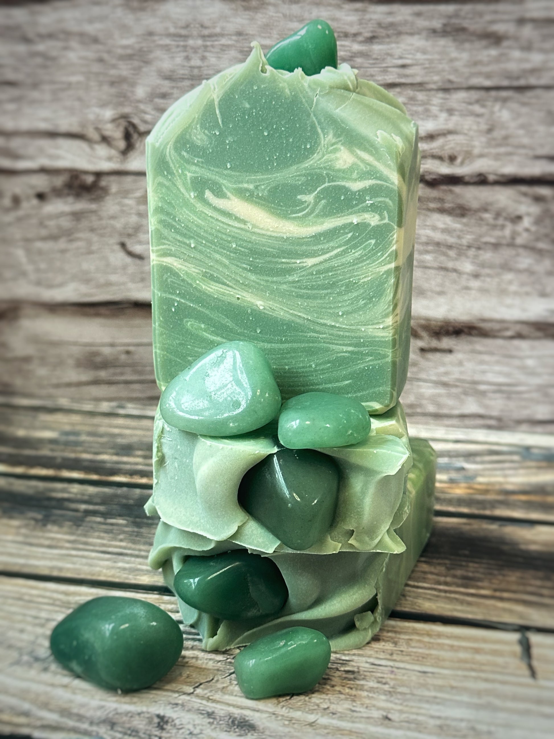 Three green artisan soap bars stacked with real green aventurine stones pressed into the tops and scattered around the base. The soap has soft swirls of mossy green and pale sage, creating a marbled, gemstone-inspired effect. The bars are set against a rustic wooden background.