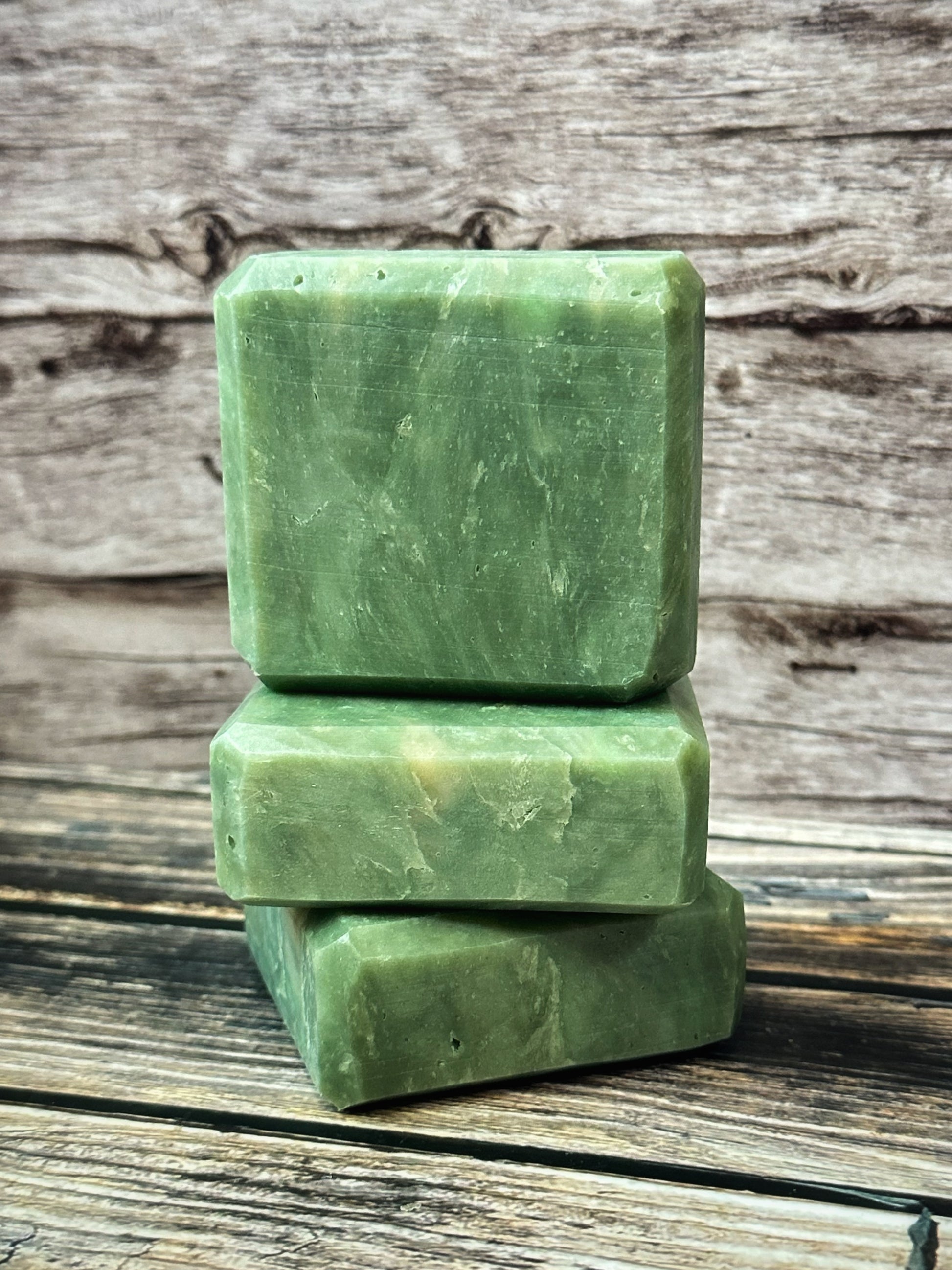 A trio of handcrafted green soap bars stacked vertically on a rustic wooden surface.  The bottom two bars are laid on their faces, one on top of the other and the third bar sits upright on top. The bars have a soft, natural marbled texture with subtle variations in tone, resembling polished jade. The background features weathered wood grain, adding an earthy, grounded aesthetic to the clean and minimalist composition.