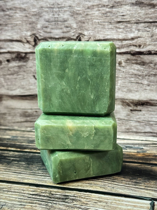 A trio of handcrafted green soap bars stacked vertically on a rustic wooden surface.  The bottom two bars are laid on their faces, one on top of the other and the third bar sits upright on top. The bars have a soft, natural marbled texture with subtle variations in tone, resembling polished jade. The background features weathered wood grain, adding an earthy, grounded aesthetic to the clean and minimalist composition.