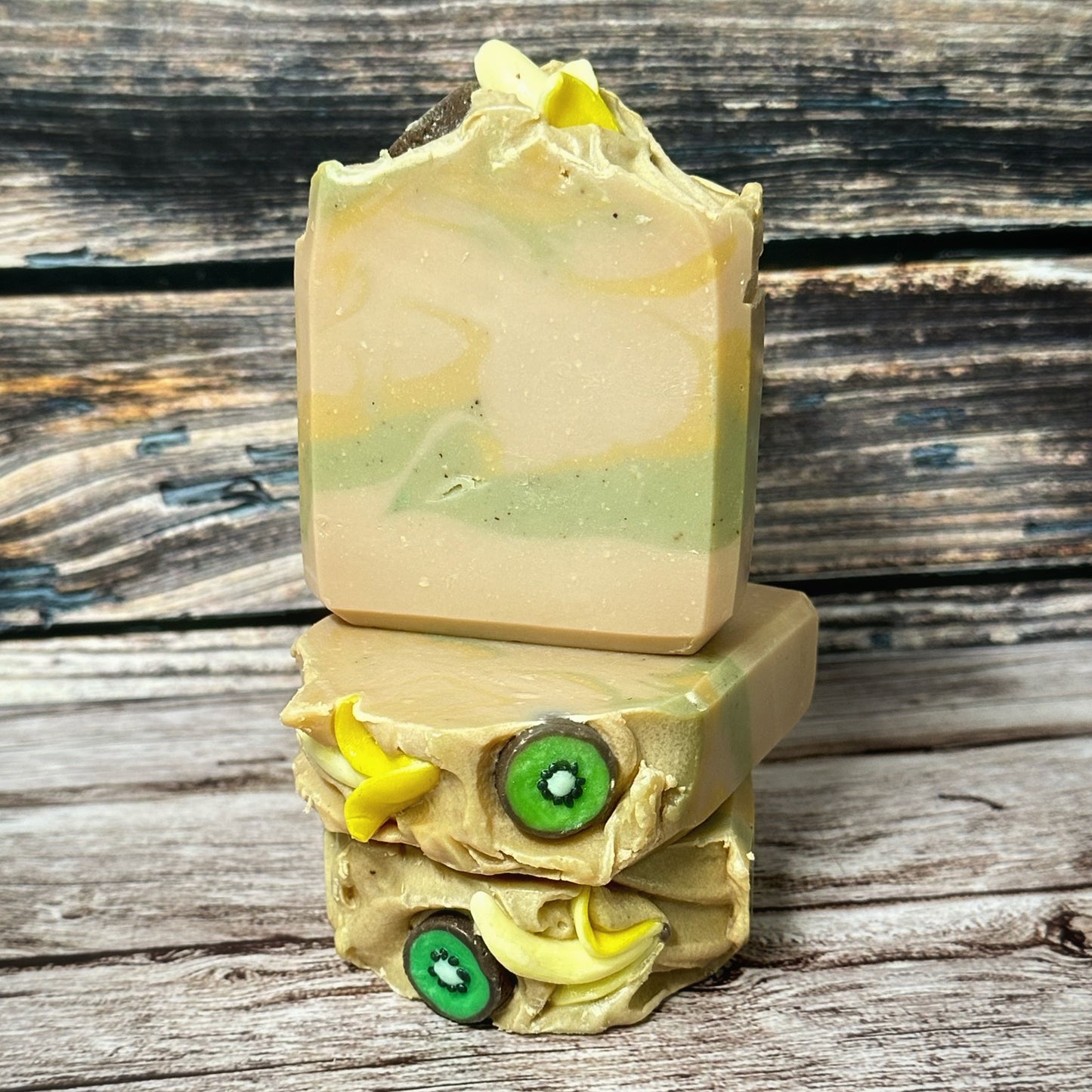 Three handcrafted soap bars stacked vertically. The front-facing bar displays soft swirls of beige, green, and light yellow. The top soap embeds resemble half peeled bananas and kiwi rounds, with a creamy textured top layer. The kiwi embeds are green with a white center and black seeds, while the banana embeds are small curved yellow pieces resembling half peeled bananas.