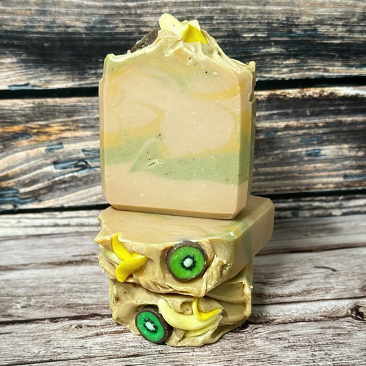 Three handcrafted soap bars stacked vertically. The front-facing bar displays soft swirls of beige, green, and light yellow. The top soap embeds resemble half peeled bananas and kiwi rounds, with a creamy textured top layer. The kiwi embeds are green with a white center and black seeds, while the banana embeds are small curved yellow pieces resembling half peeled bananas.
