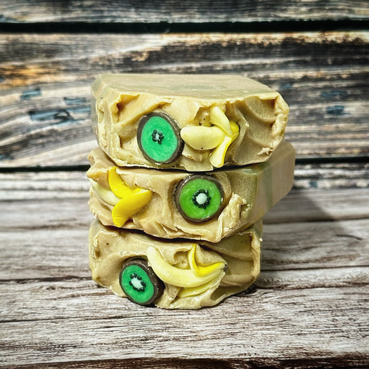 Three stacked soap bars showcasing detailed embed work. Each bar features bright green kiwi slices with black seeds and small, curved yellow banana embeds nestled into a textured tan frosting-like top. The embed fruits are three-dimensional and are made from soap.