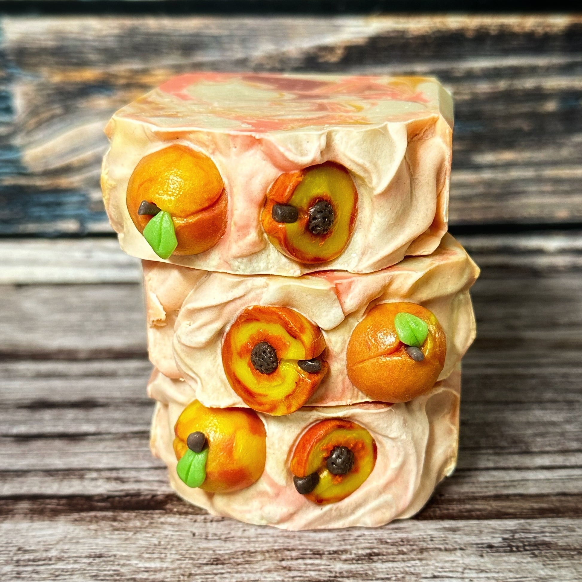 Close-up of three handmade soap bars stacked to show their tops. Each soap is topped with realistic peach-themed embeds, including whole and halved peaches with green leaves and dark brown pits. The tops have a textured swirl design in peach and cream tones.