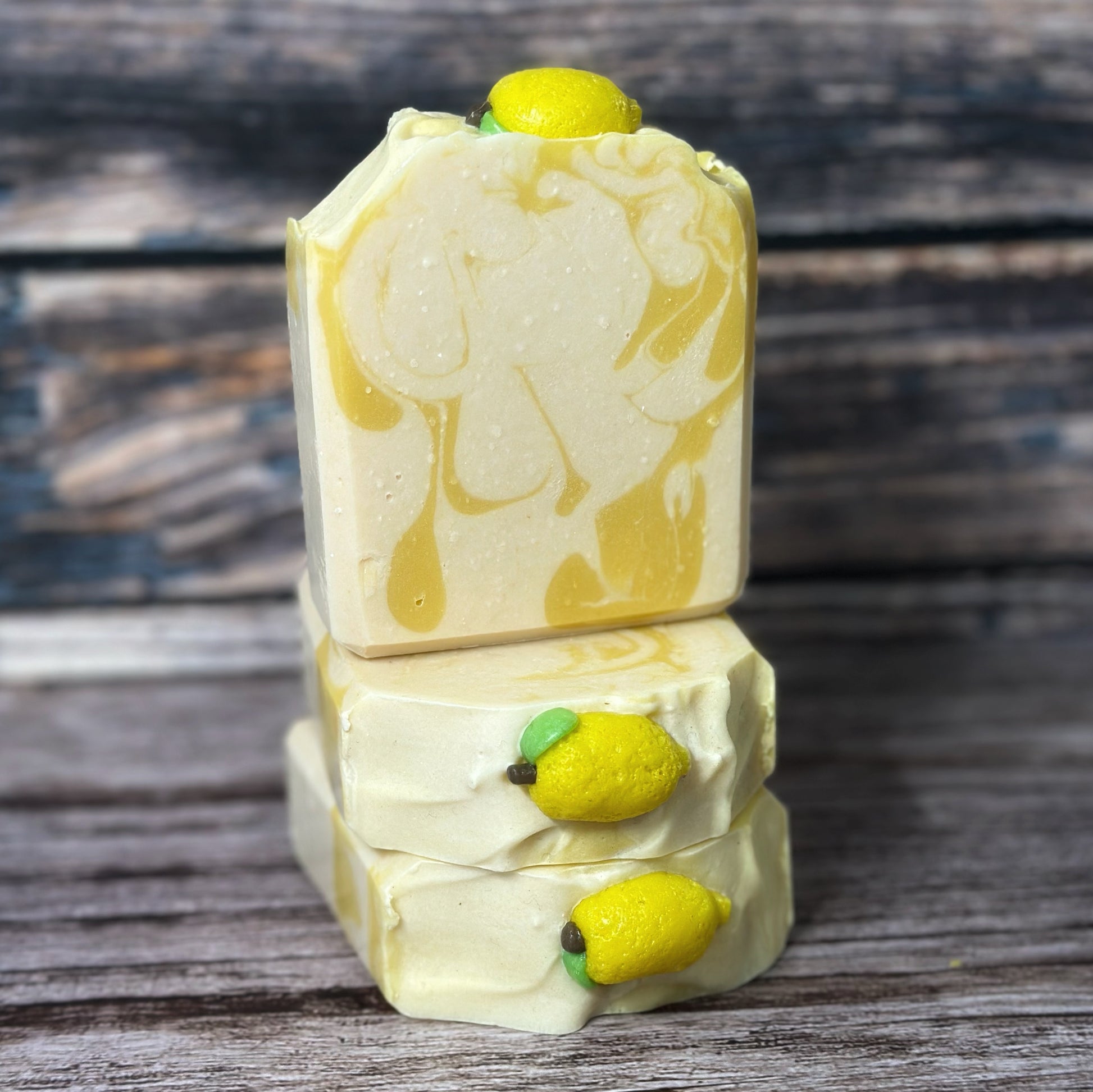 Three handmade lemon soaps stacked on a rustic wood background. Each bar features a yellow and cream swirl design and is topped with a small, realistic lemon-shaped soap embed.