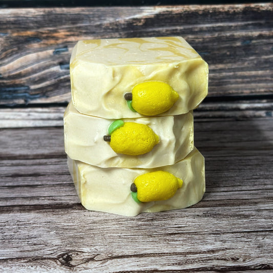 Close-up view of three stacked lemon-themed soap bars, showcasing the tops of the bars. Each top features a bright yellow lemon-shaped soap embed with a green leaf and brown stem, embedded into a pale yellow swirled surface. Set against a rustic wood background.