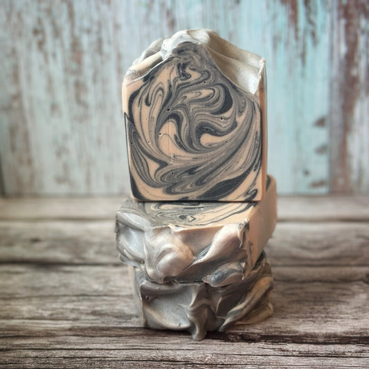A handcrafted bar of soap with a striking black and pink swirl design, resembling marble. The top is sculpted into flowing peaks. The soap is displayed stacked in a trio - one standing upright and two laid flat - on a rustic wood surface with a weathered teal and brown wood backdrop.