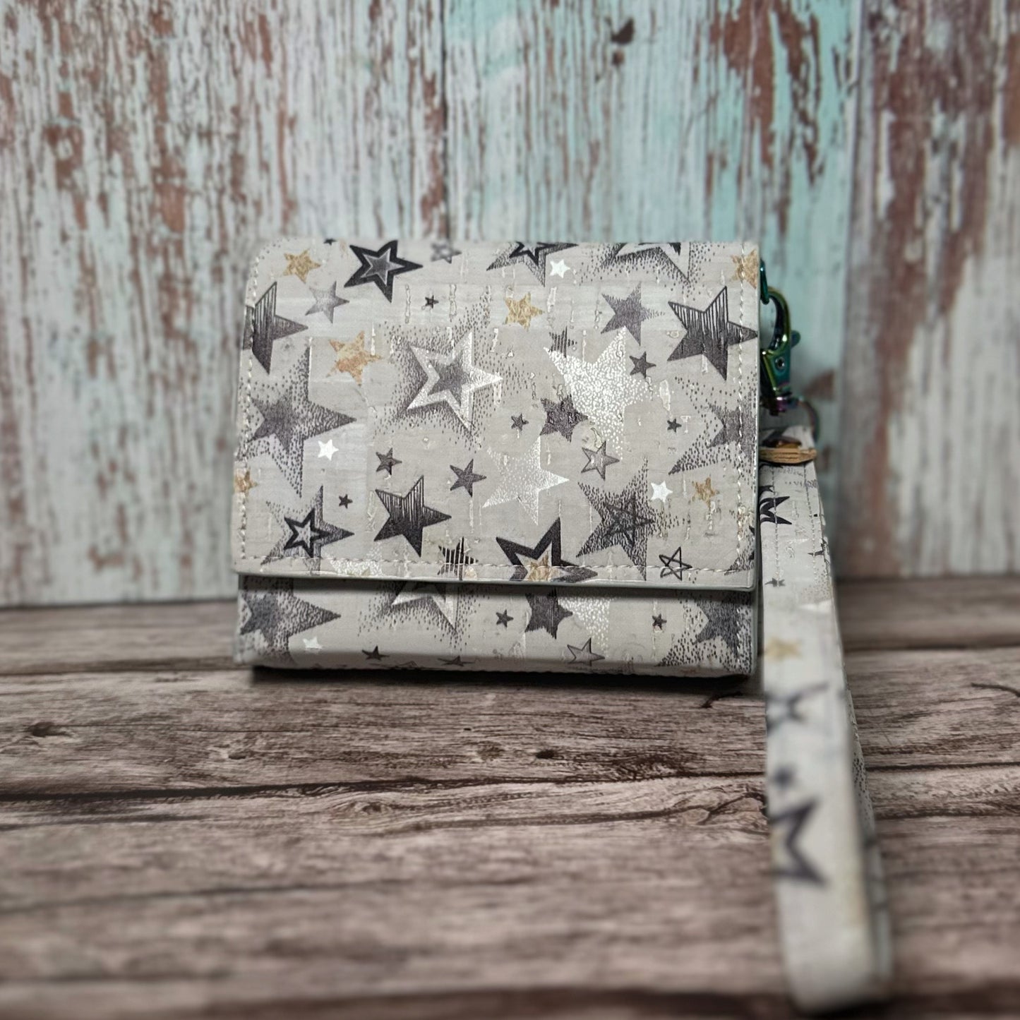 Front view of a handmade wallet crafted from light-colored cork material with a scattered star print in shades of black, silver, and gold. The wallet is shown closed with a matching star-print wrist strap attached. It rests on a rustic wooden surface with a distressed wood backdrop in soft teal and brown tones.