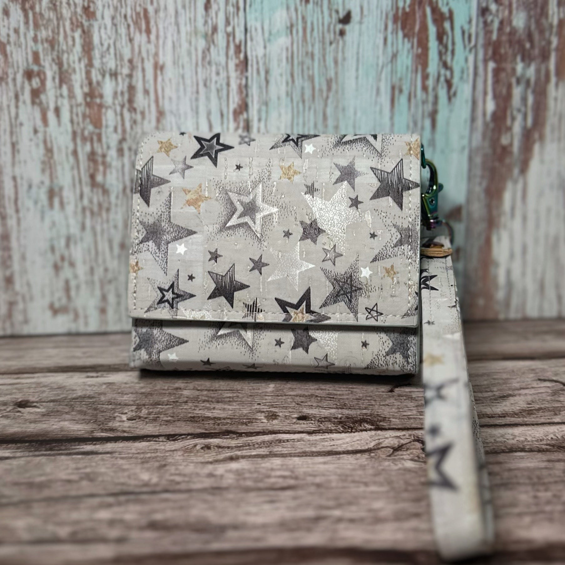 Front view of a handmade wallet crafted from light-colored cork material with a scattered star print in shades of black, silver, and gold. The wallet is shown closed with a matching star-print wrist strap attached. It rests on a rustic wooden surface with a distressed wood backdrop in soft teal and brown tones.