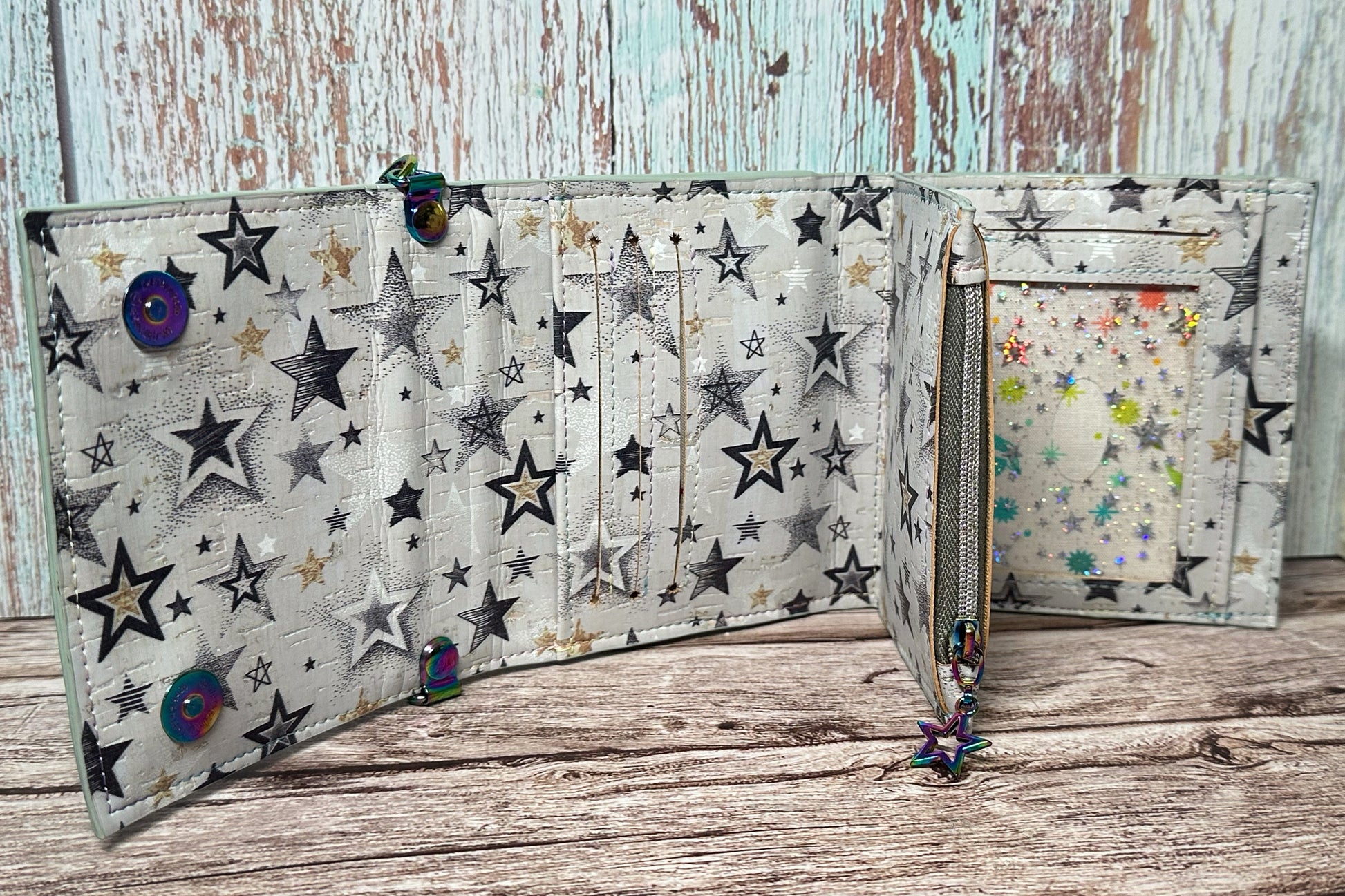 Interior view of a handmade wallet crafted from light cork with a metallic black, silver, and gold star pattern. The wallet features three horizontal card slots, a cash pocket behind them, a center zippered coin pouch with a rainbow star zipper pull, and a starry holographic ID window with a multi-color insert behind it. The lining matches the outer cork material. The wallet is displayed open on a rustic wood surface with a distressed teal and brown wood backdrop.