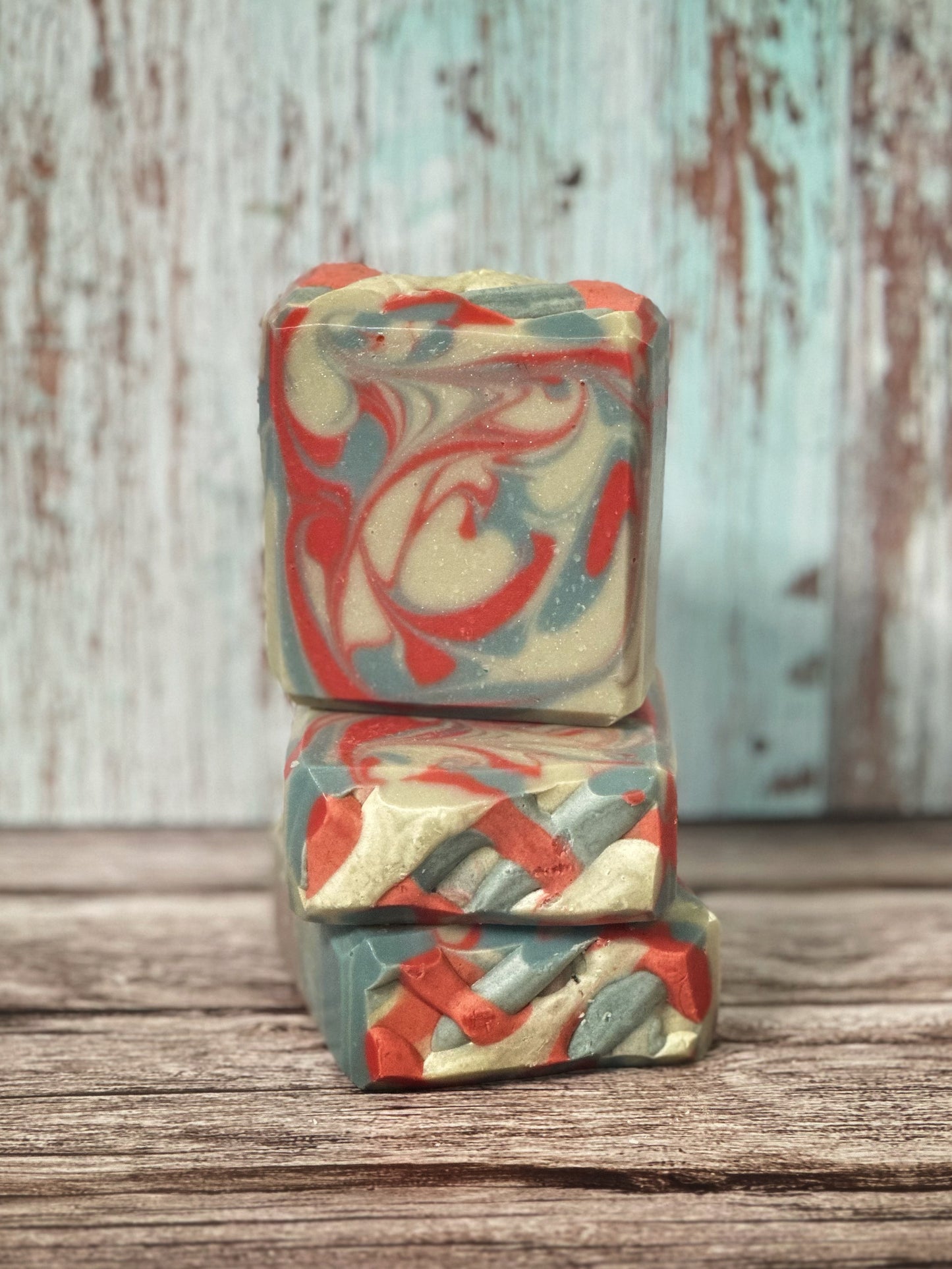 Three handcrafted soap bars are arranged in a vertical stack on a rustic wooden surface. Two bars are laid flat, and a third sits upright on top. The soap features a swirling design in red, slate blue, and off-white, creating a marbled effect across all sides. The background is made up of weathered vertical wood planks in pale aqua and warm brown tones.