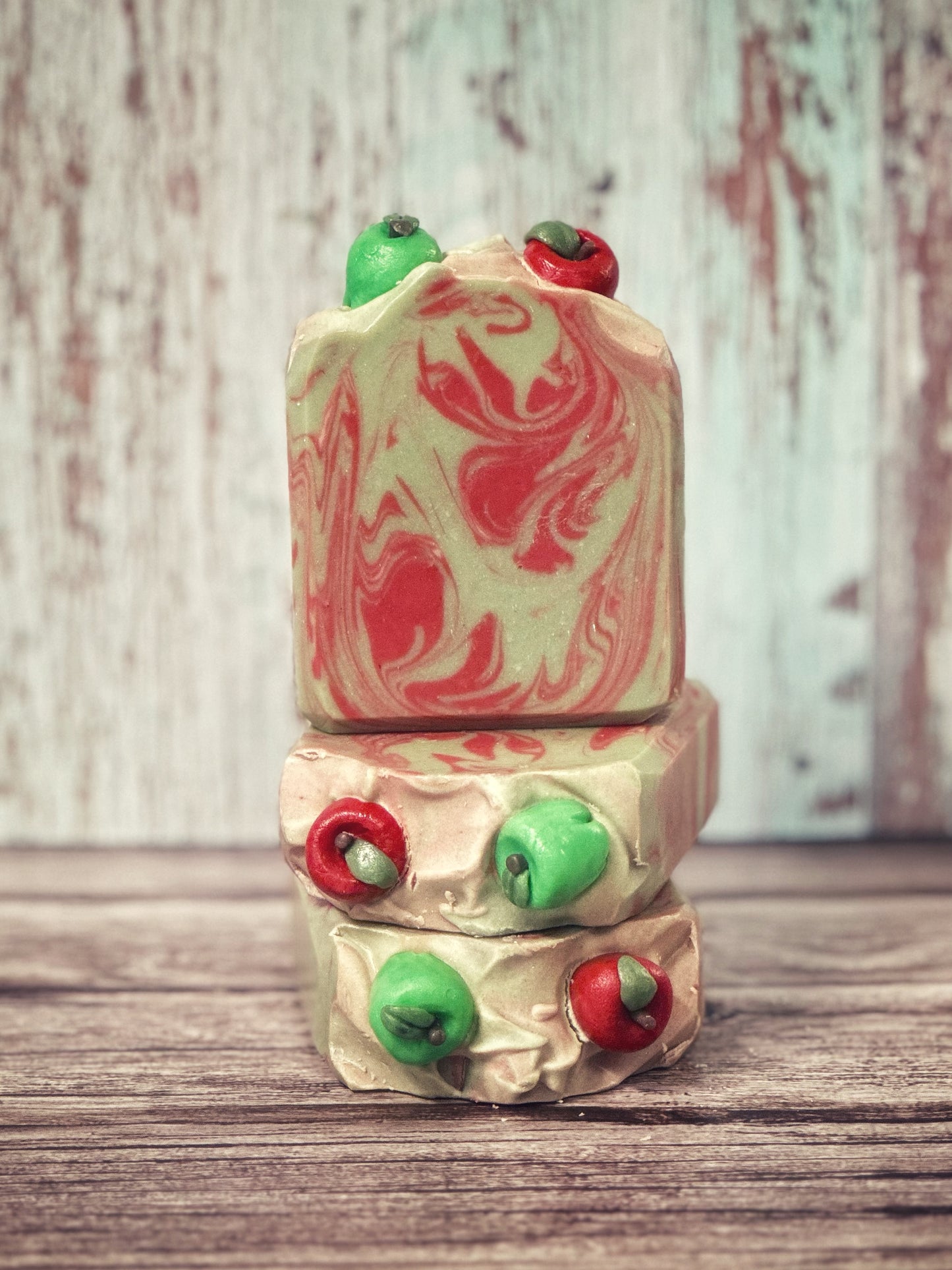 Three bars of handcrafted soap are arranged in a vertical stack on a wooden surface. Two bars lie flat, one on top of the other, and a third bar stands upright, centered on top. The soap features a bold swirl pattern in bright red and creamy pale green. Each bar is topped with three-dimensional sculpted apples in red and green. The background is a distressed light wood panel.